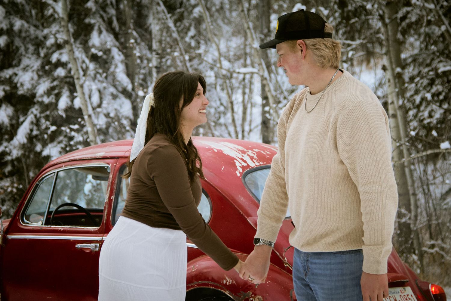 Couple holding hands by a red car in a snowy winter forest setting.