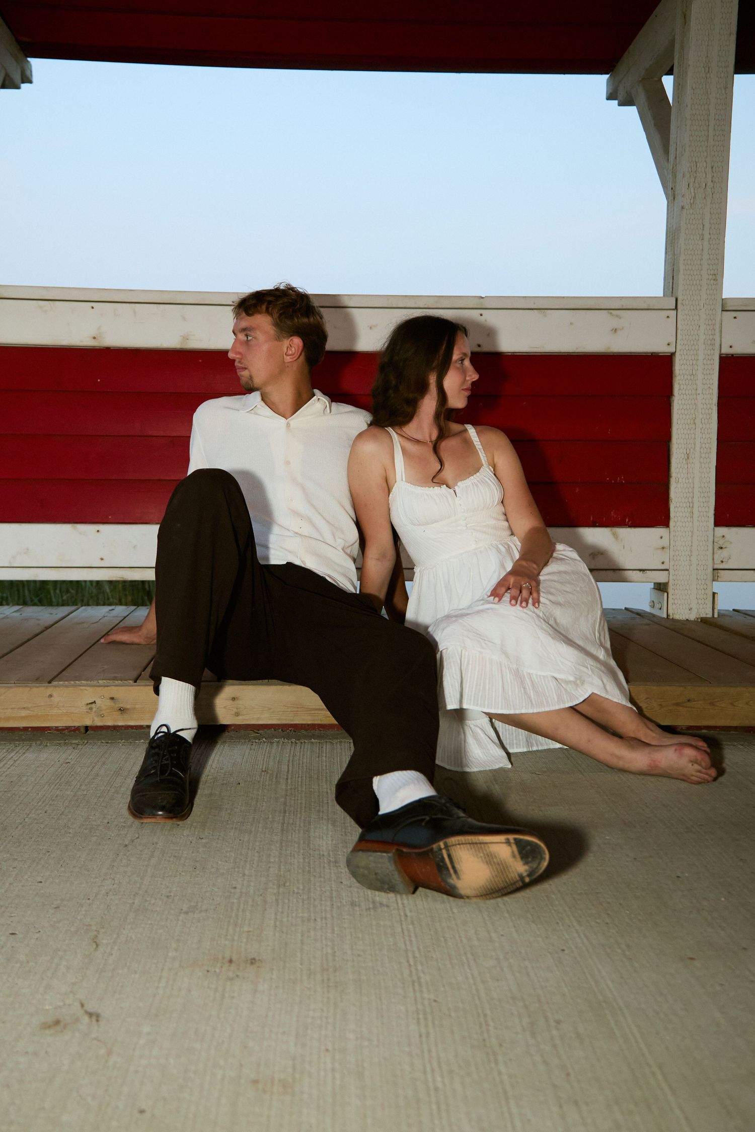 A young couple sits back-to-back on a bench under a red and white covered porch, appearing distant.