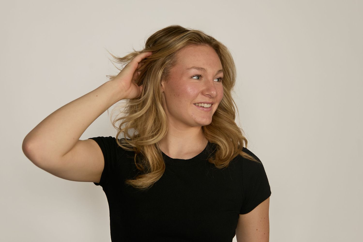 A person with wavy blonde hair wearing a black t-shirt poses against a neutral background while touching their hair.