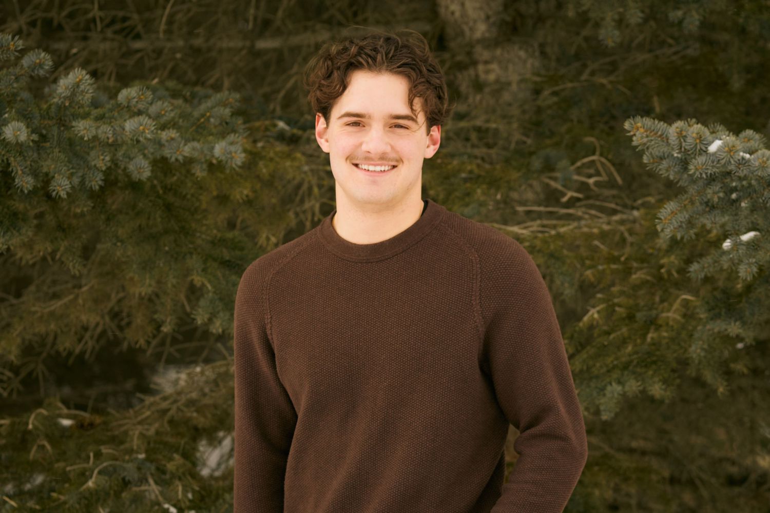 A person in a brown sweater smiles while standing outdoors against a backdrop of green foliage.