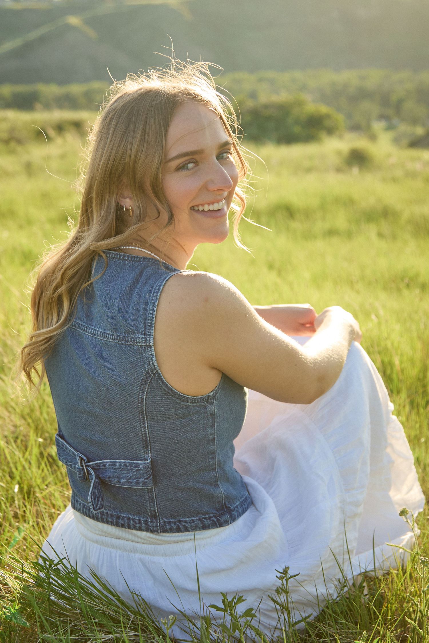 Young blonde woman in denim vest and white skirt sitting in green meadow, smiling over shoulder in golden sunlight.