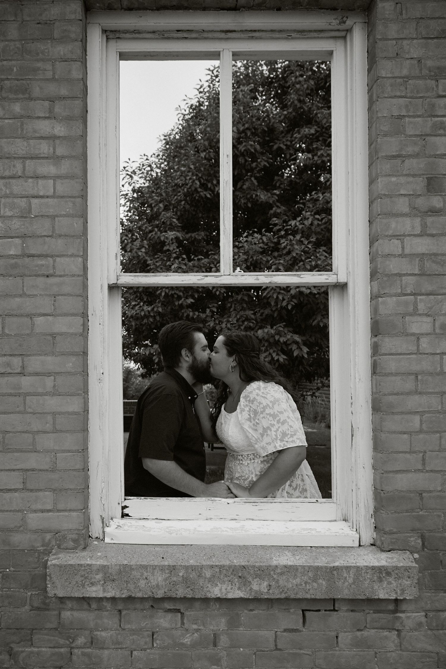 Black and white photo of a couple kissing framed in a vintage window of a brick building with trees in background.