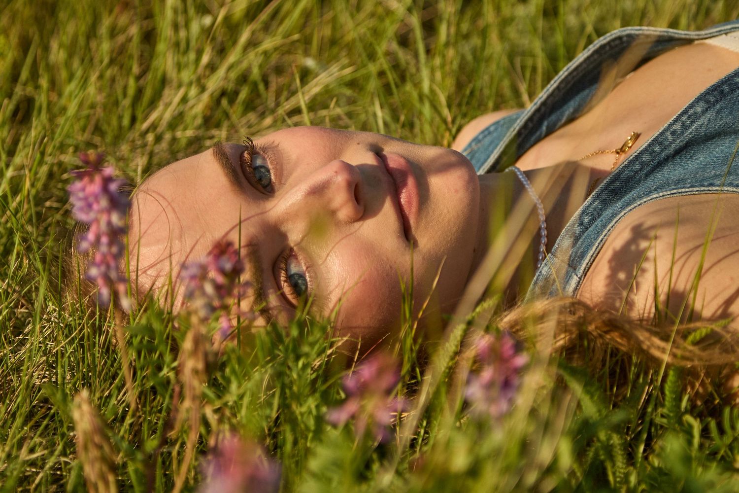 Young woman lying in green grass field with wildflowers, bathed in warm golden sunlight, serene outdoor portrait.