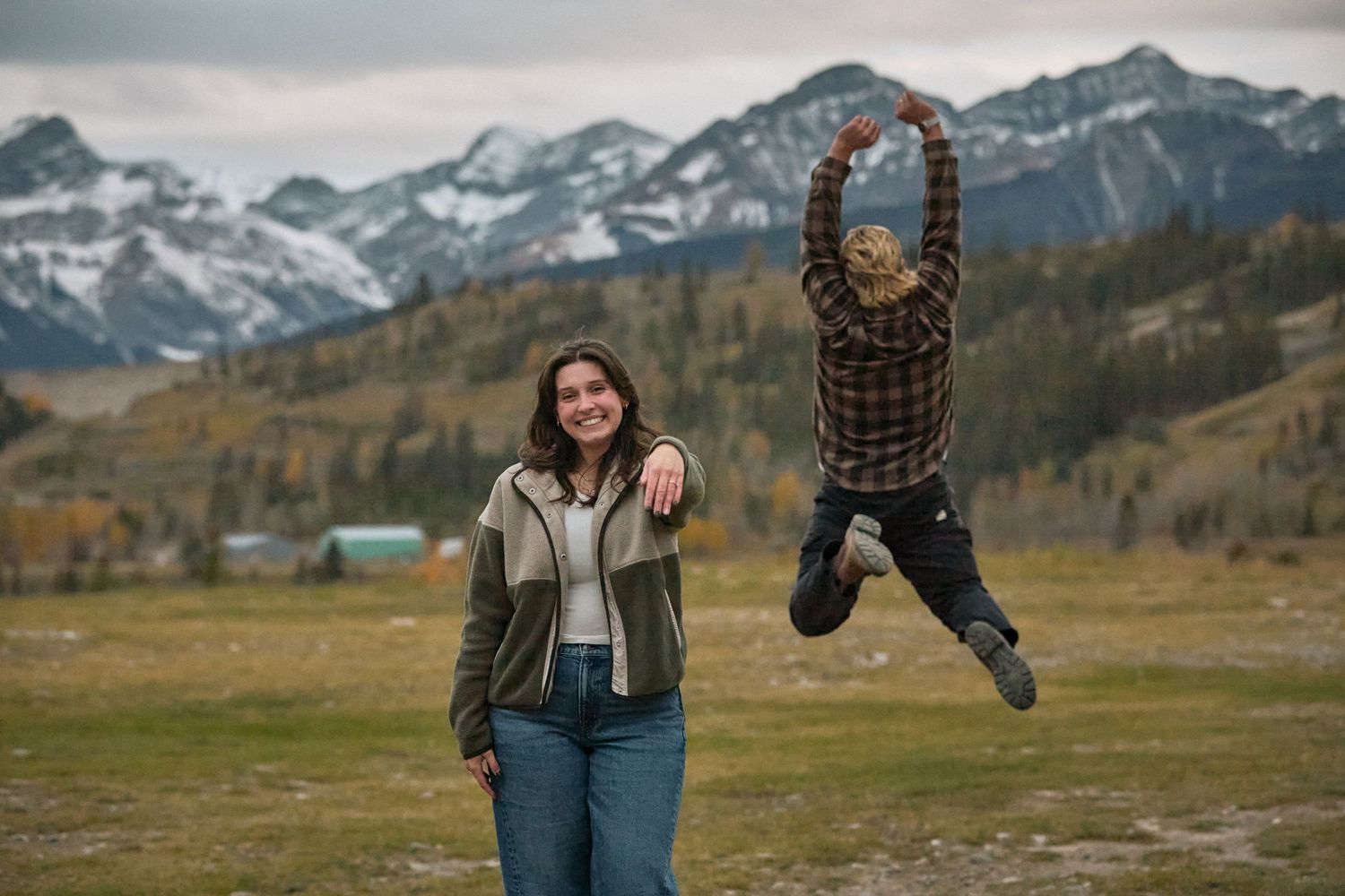 Two people pose in a mountain meadow, one jumping joyfully, with snow-capped peaks and autumn trees in the background.