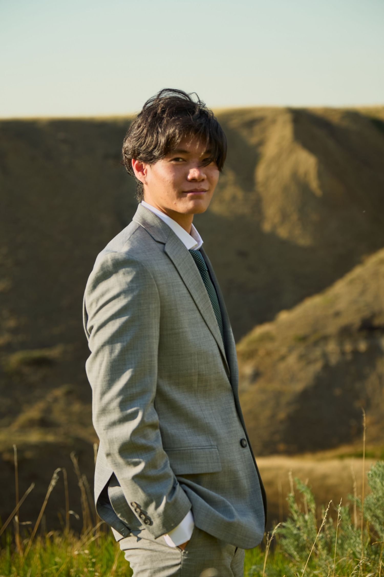 Highschool graduate in a gray suit poses against a scenic mountain backdrop at golden hour.
