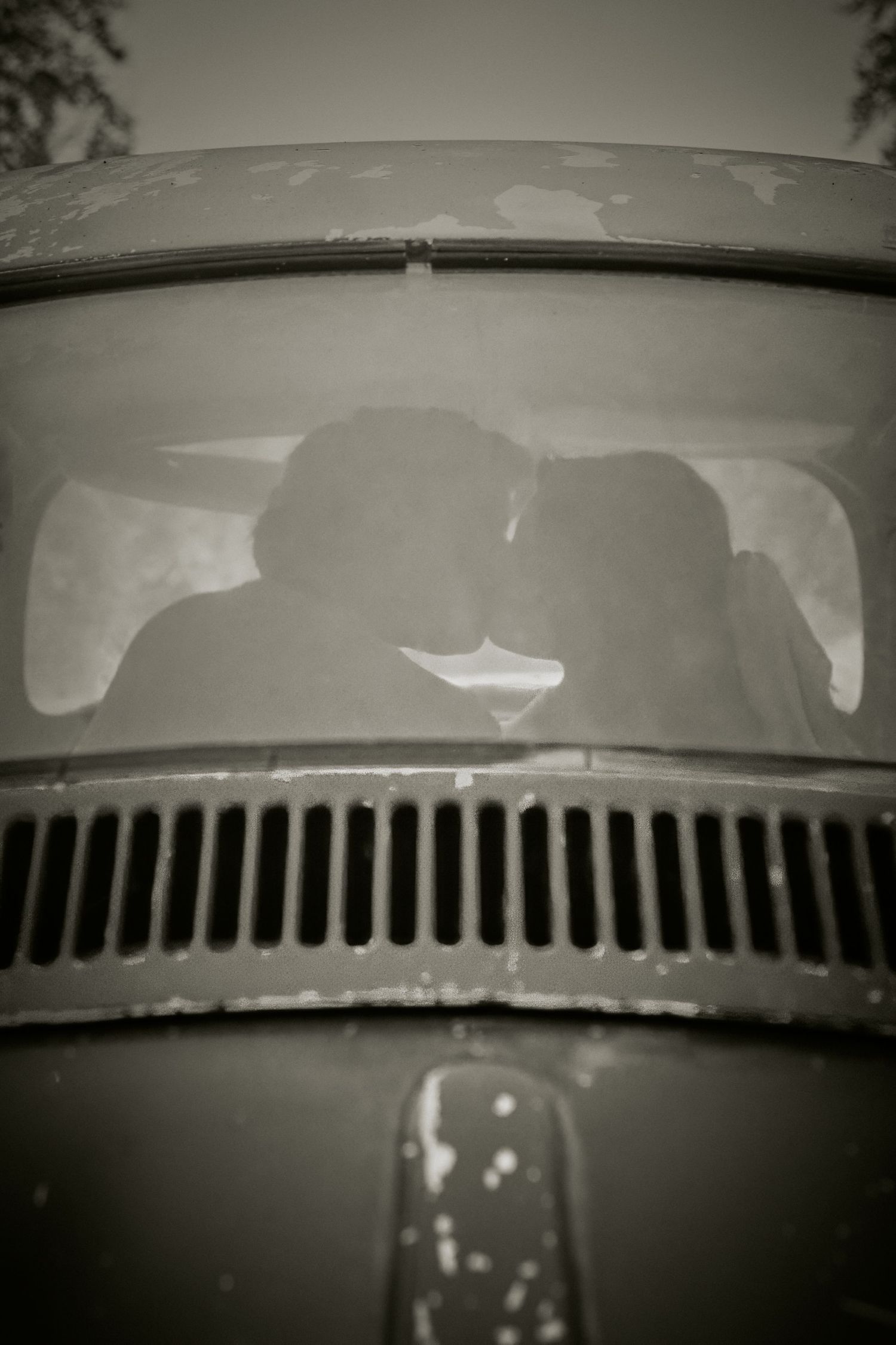 Silhouettes visible through windshield of vintage car with distinctive front grille in black and white photograph.