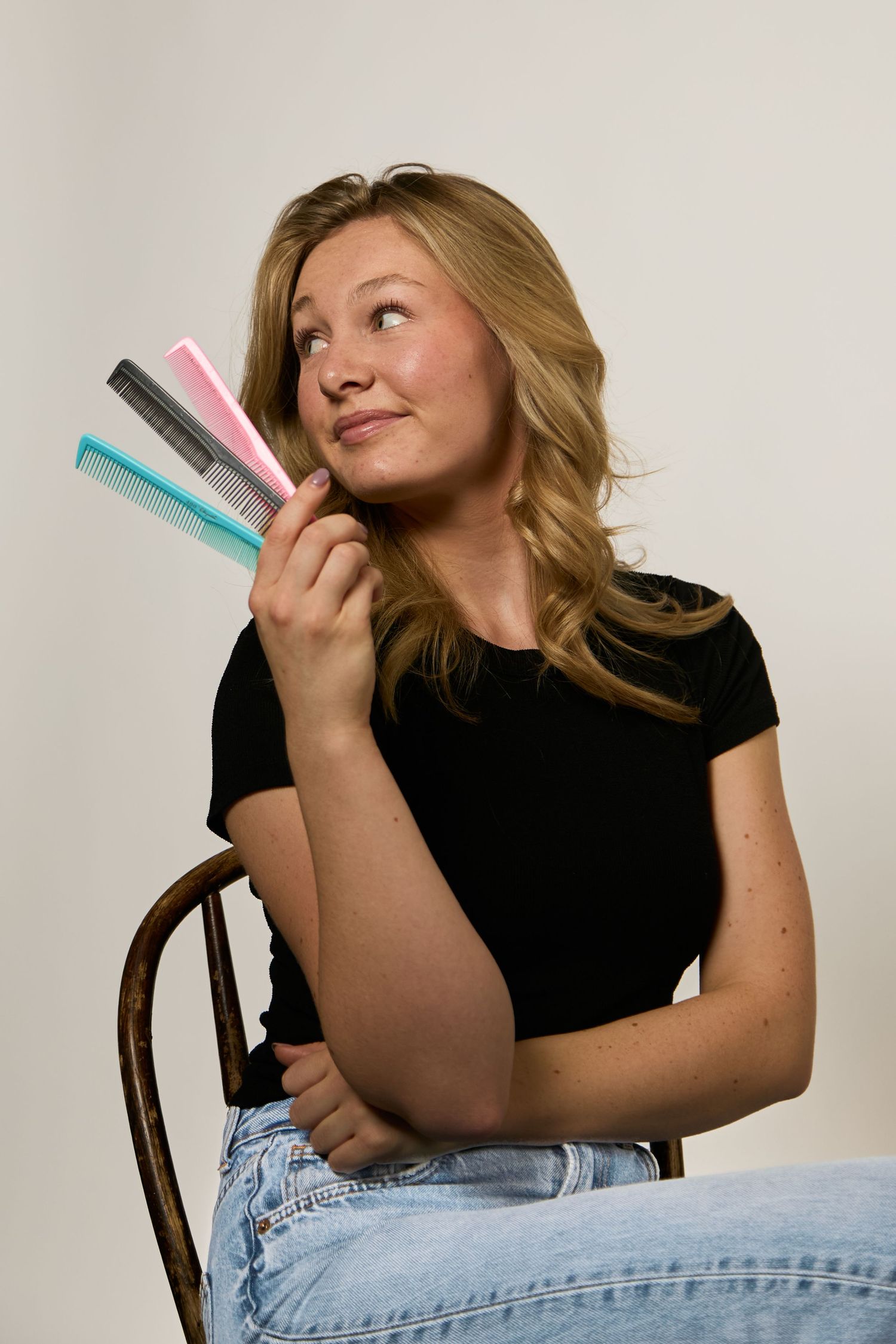 A smiling blonde woman in a black top and jeans sits on a chair, holding colorful hair combs while looking upward.