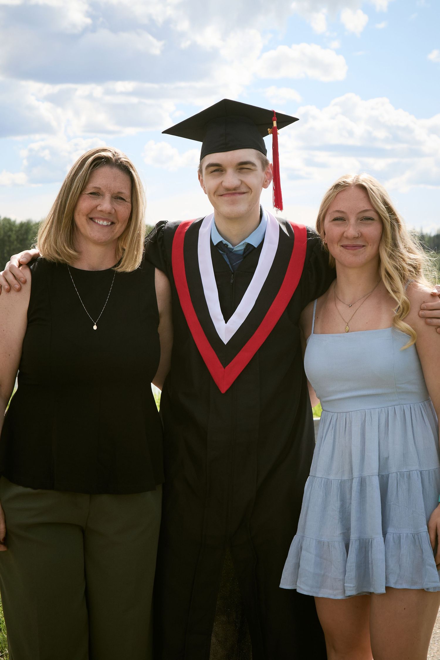 A young man in graduation cap and gown smiles with two women outdoors under a partly cloudy sky.