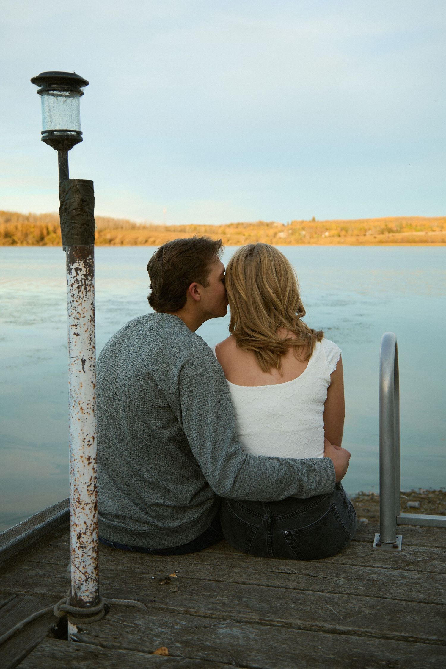 A couple sits on a wooden dock kissing beside a rustic lamp post with a calm lake and autumn trees in the background.