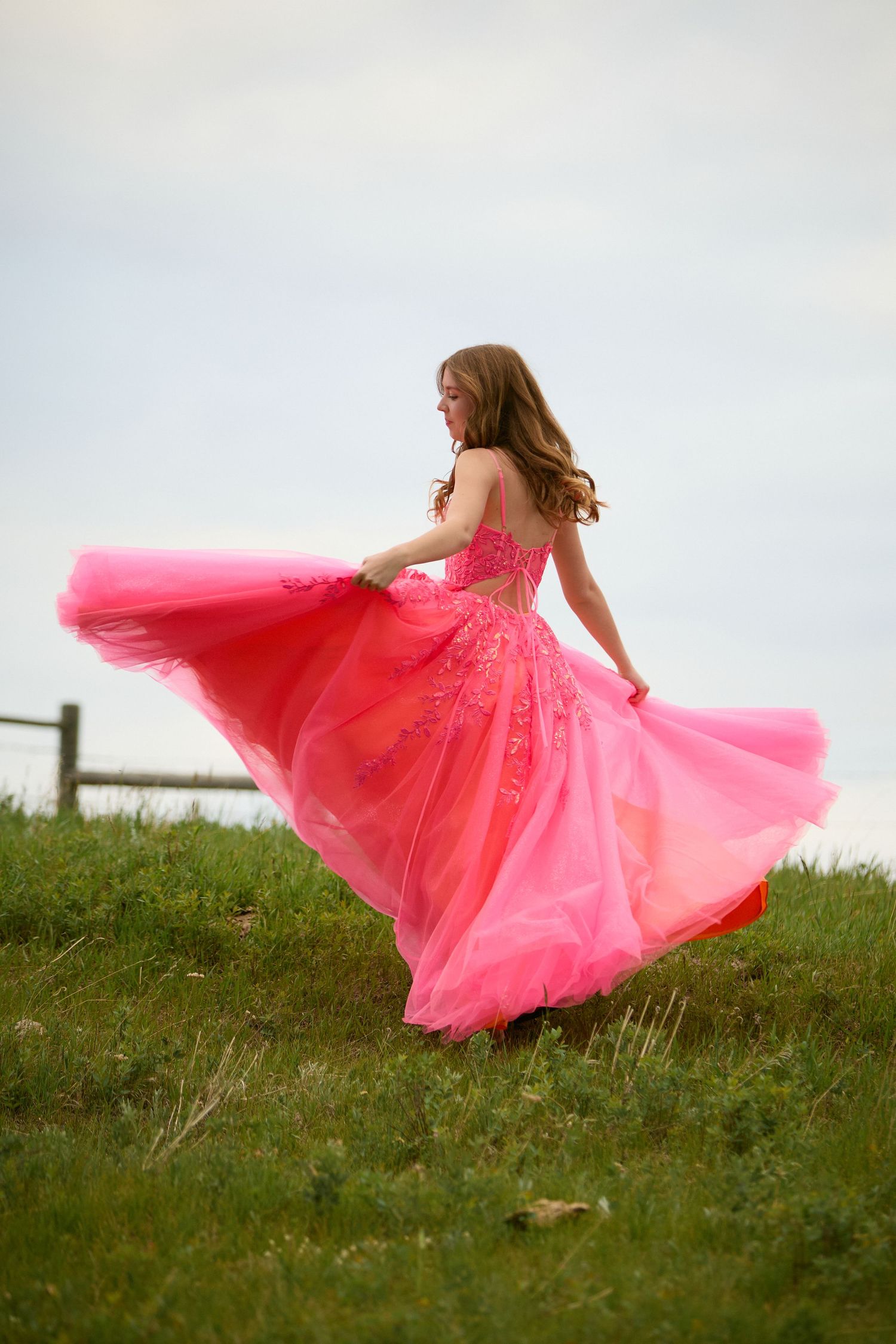 Young woman twirling in a flowing pink ball gown on a green hillside, with a wooden fence in the background.