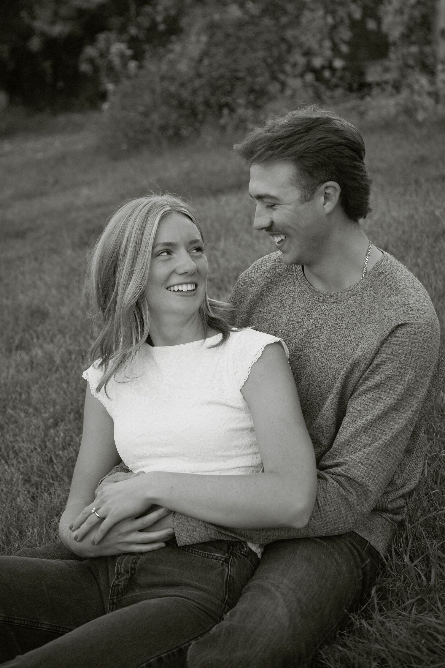 Happy couple sitting on grass, smiling at each other in a romantic black and white outdoor portrait photo.