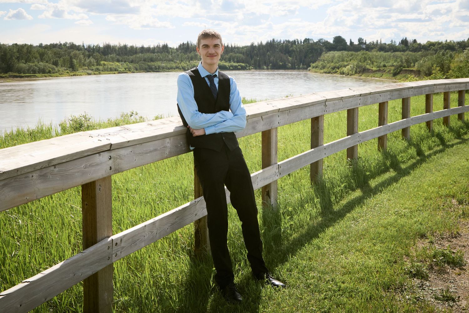Young man in formal attire with crossed arms leaning on wooden fence by a scenic river and green landscape.