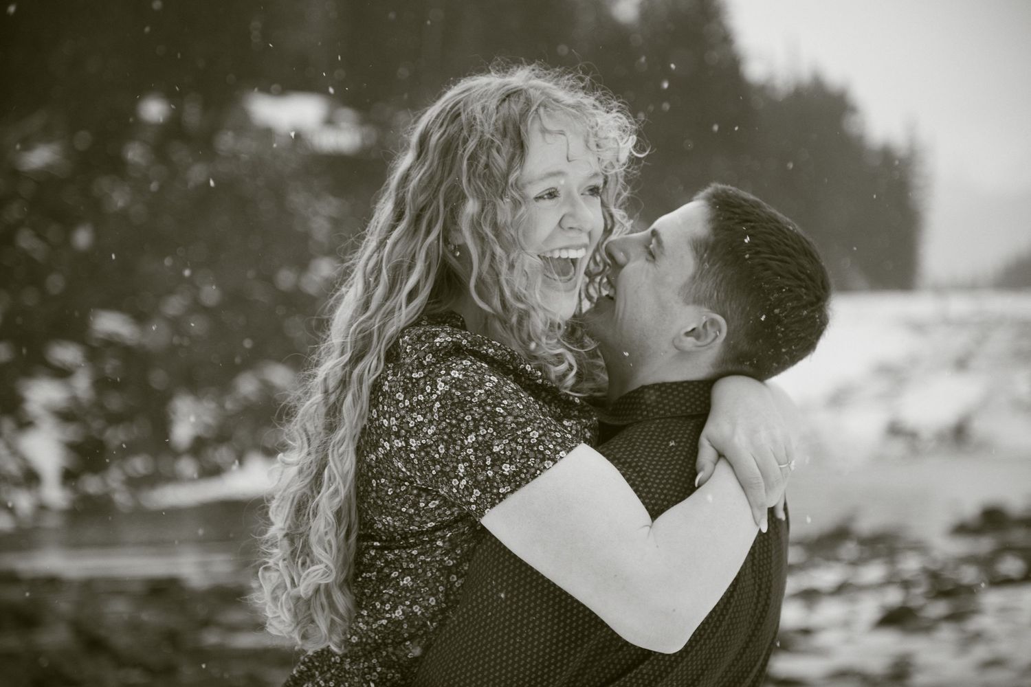 Black and white photo of a joyful couple embracing in snowy winter scenery, woman laughing as man kisses her cheek.