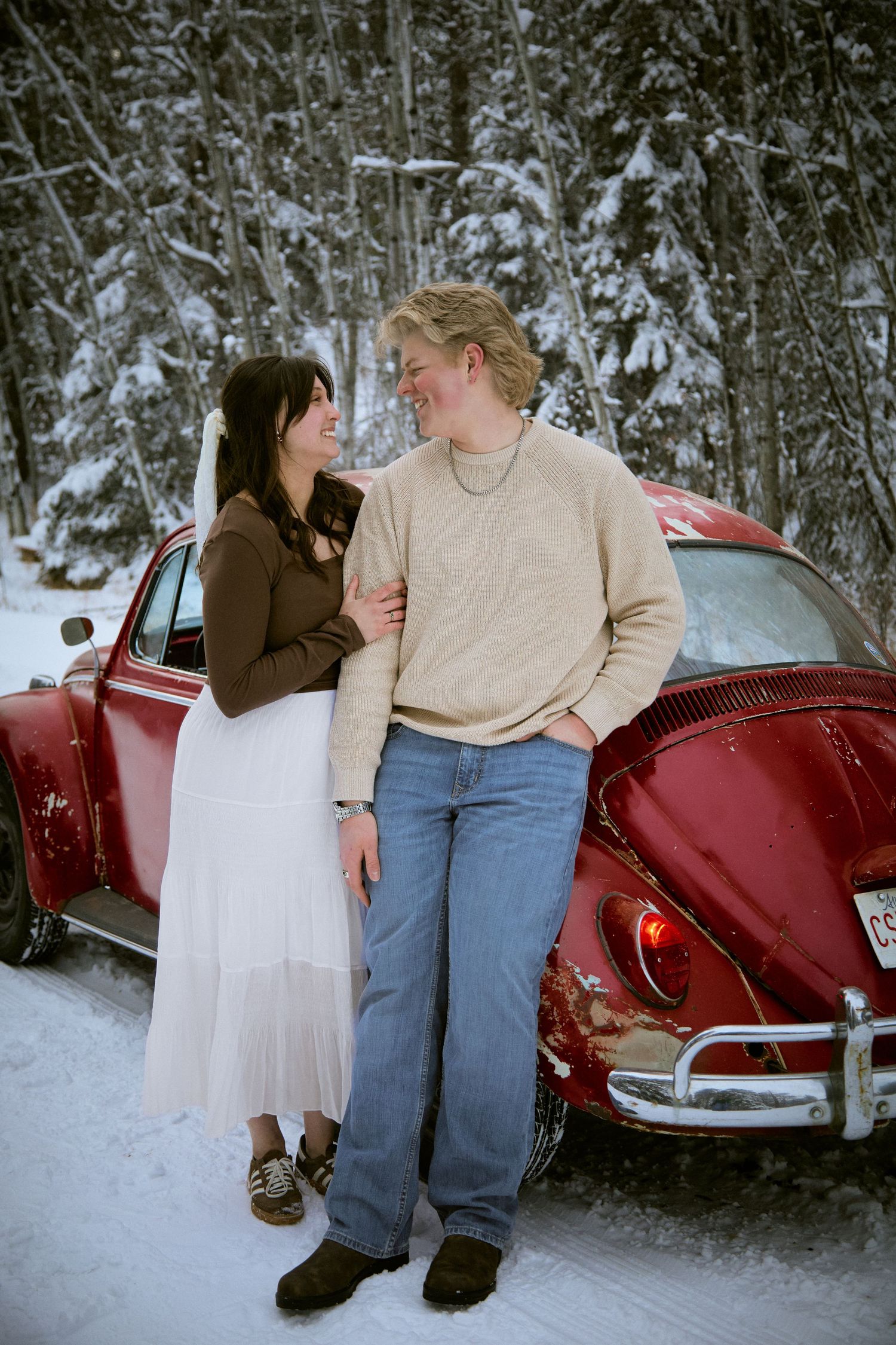 Couple smiling at each other in winter, leaning against a vintage red VW Beetle surrounded by snow-covered trees.