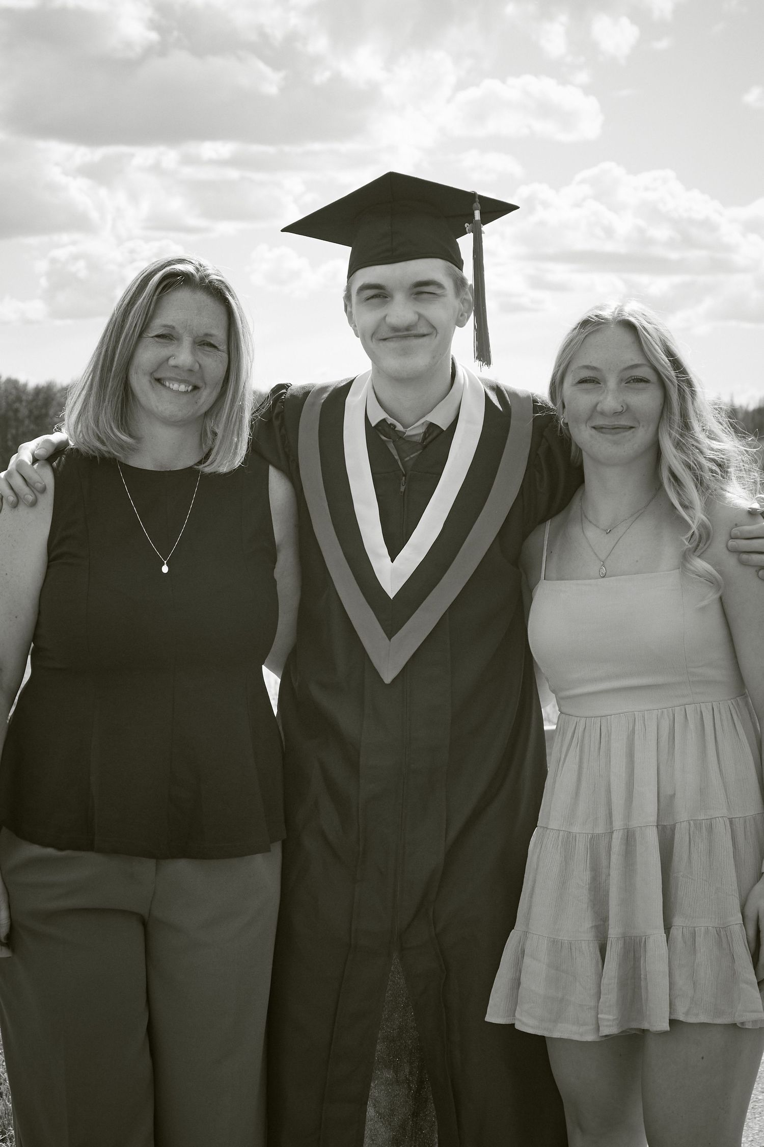 Black and white photo of a smiling graduate in cap and gown posing with two women outdoors on graduation day.