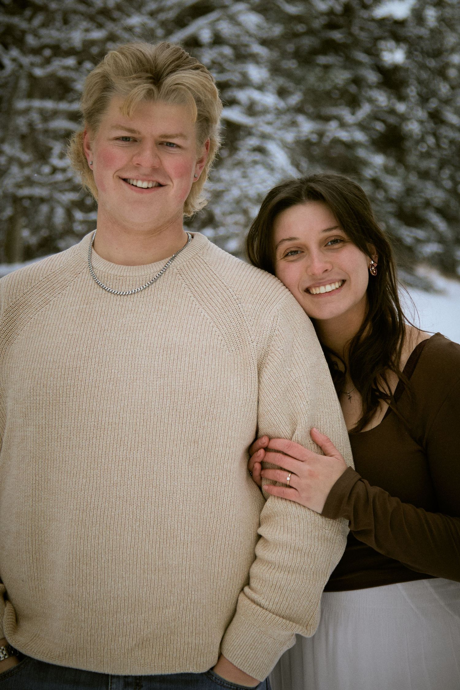 A couple poses together in a winter setting wearing a beige sweater and dark clothing against a snowy background.