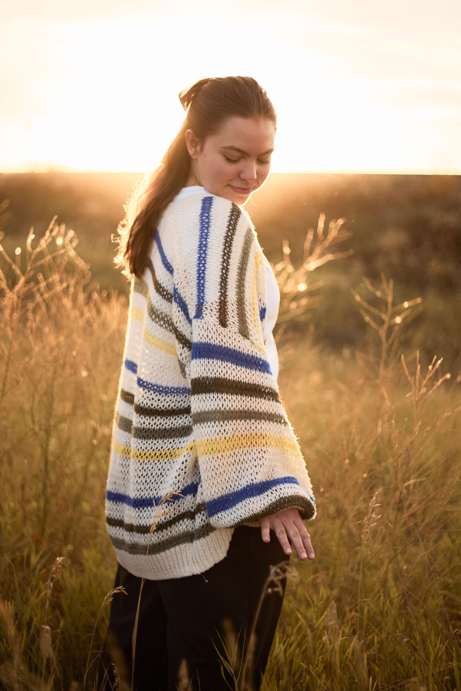 A person wearing a striped blue and white sweater stands in a golden field at sunset, creating a warm autumn scene.