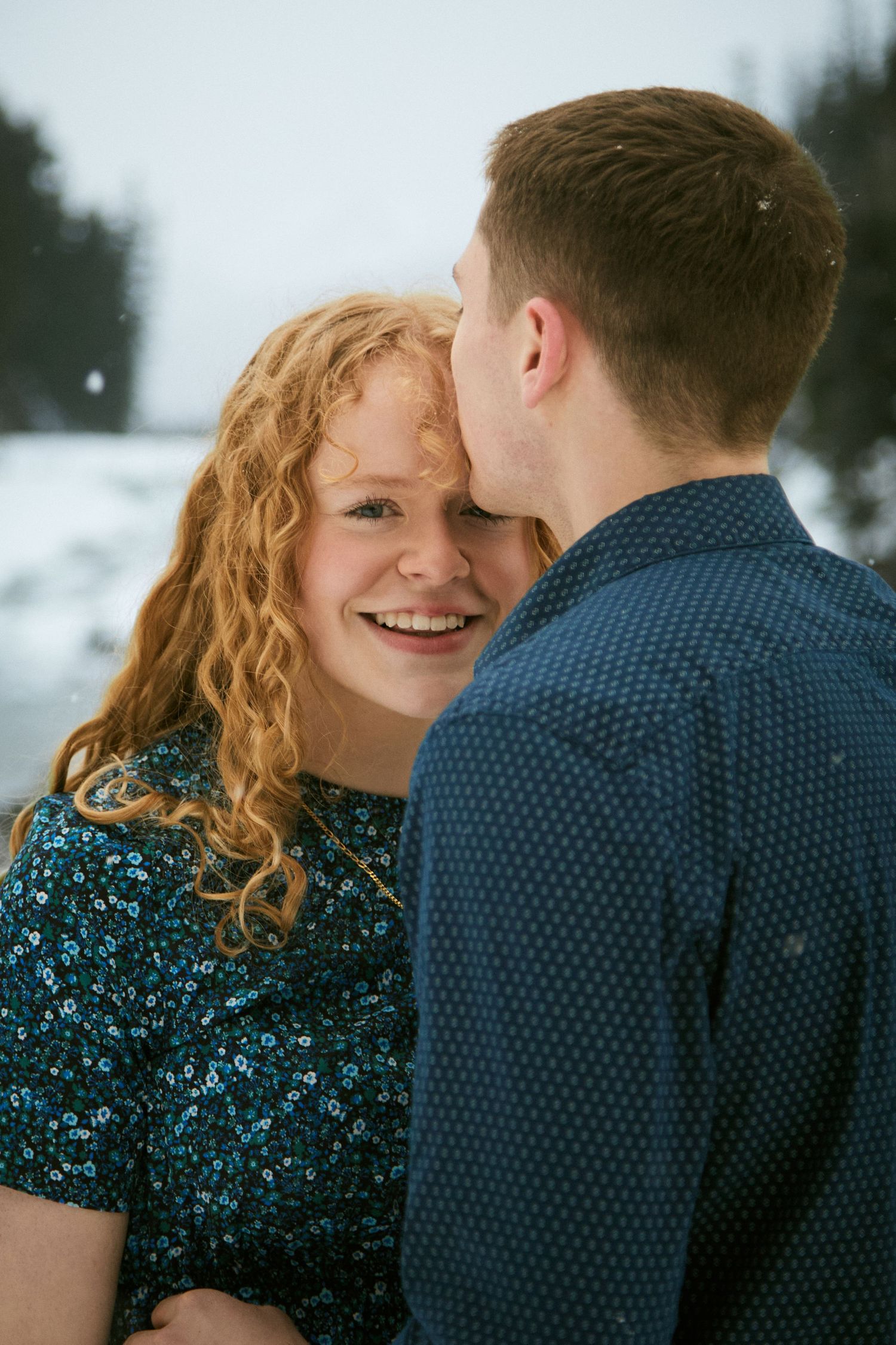 A smiling redhead woman receives a kiss on the cheek from a man outdoors in a snowy winter setting.