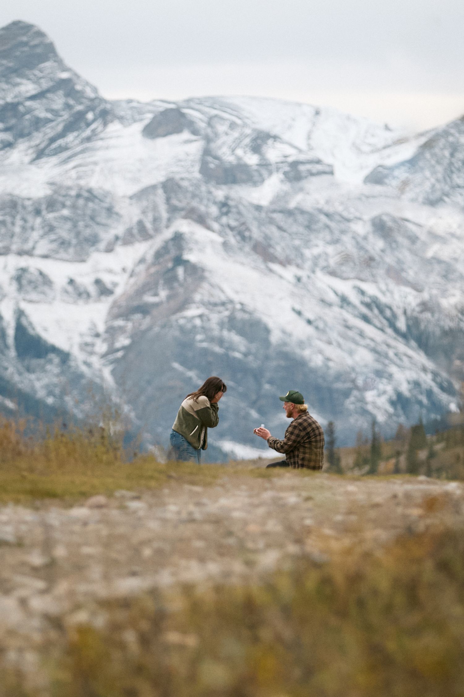 Couple sharing a romantic proposal moment on a mountain trail with snow-capped peaks in the background.