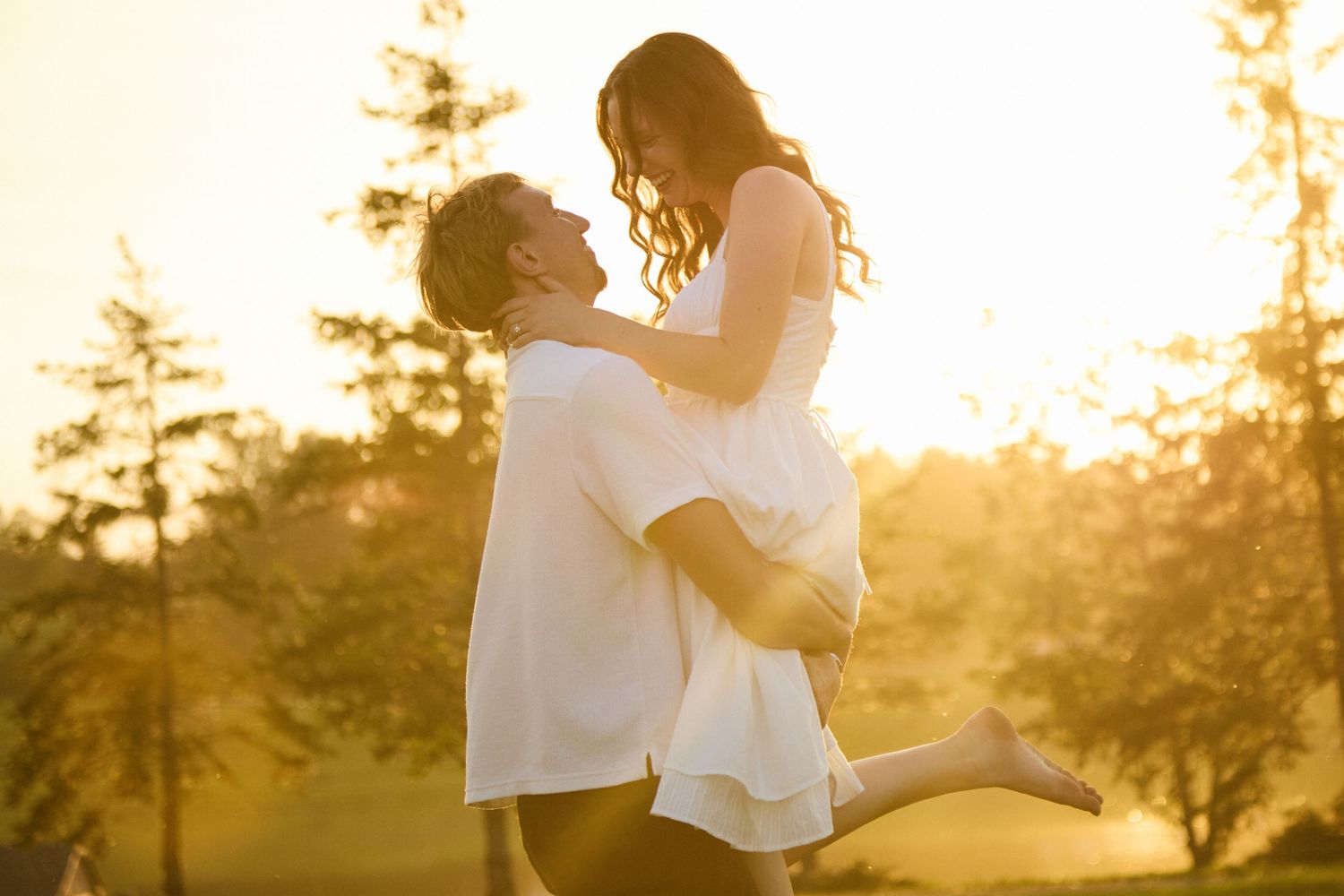 Couple sharing a romantic moment at golden hour, man lifting woman in white dress outdoors.