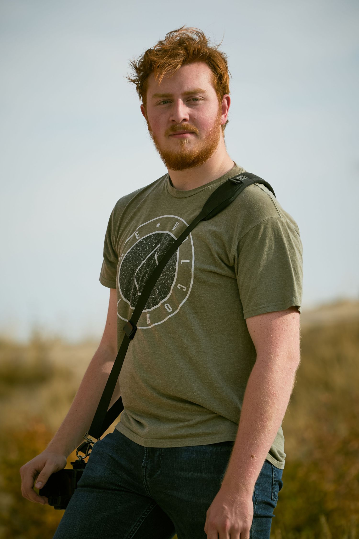 Photographer in olive t-shirt with graphic design poses outdoors in natural lighting.