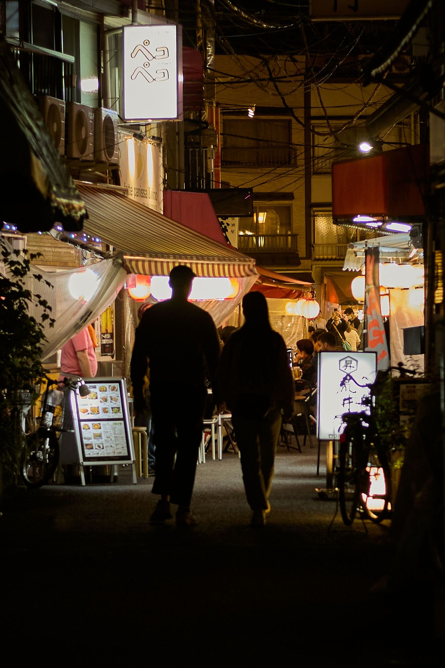 Two silhouetted figures walk through a glowing Japanese alley at night, lined with lanterns and restaurant signs.