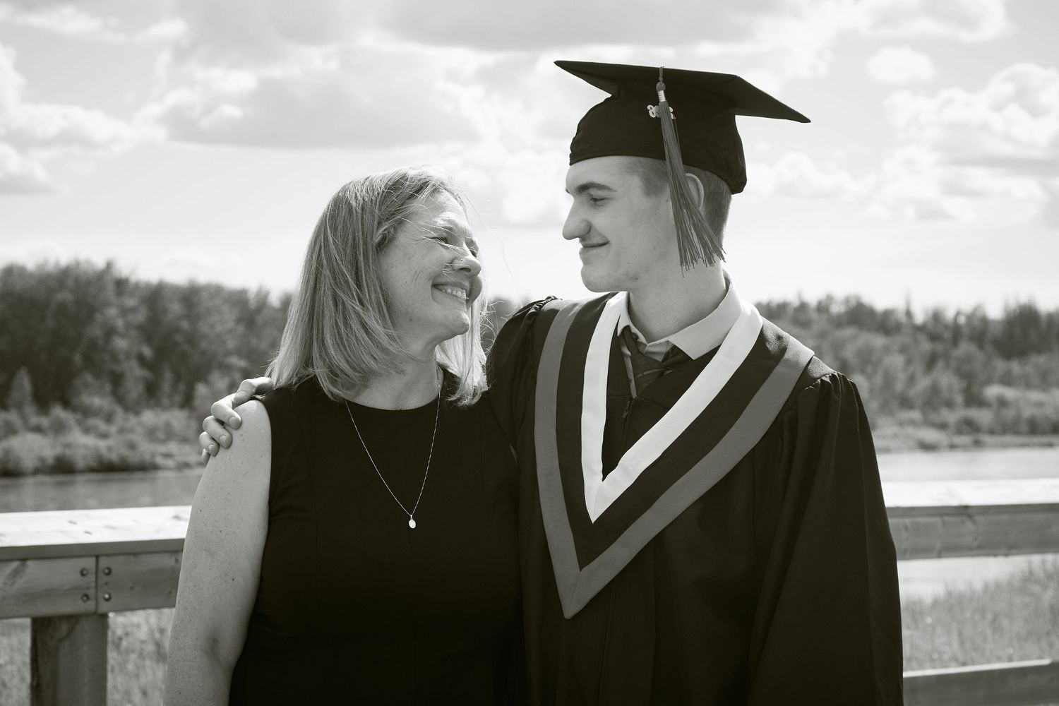 A graduate in cap and gown smiles with mom on a bridge, captured in black and white.