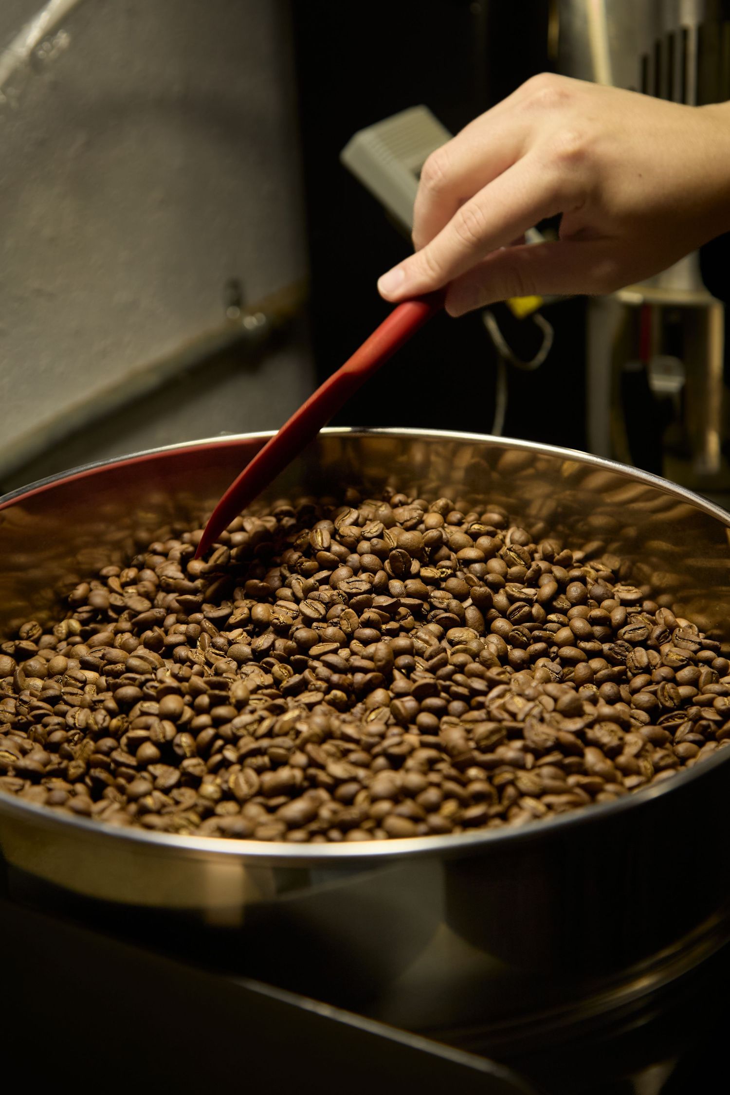 Coffee beans being roasted in a dark metal pan with a red-handled spoon stirring them.