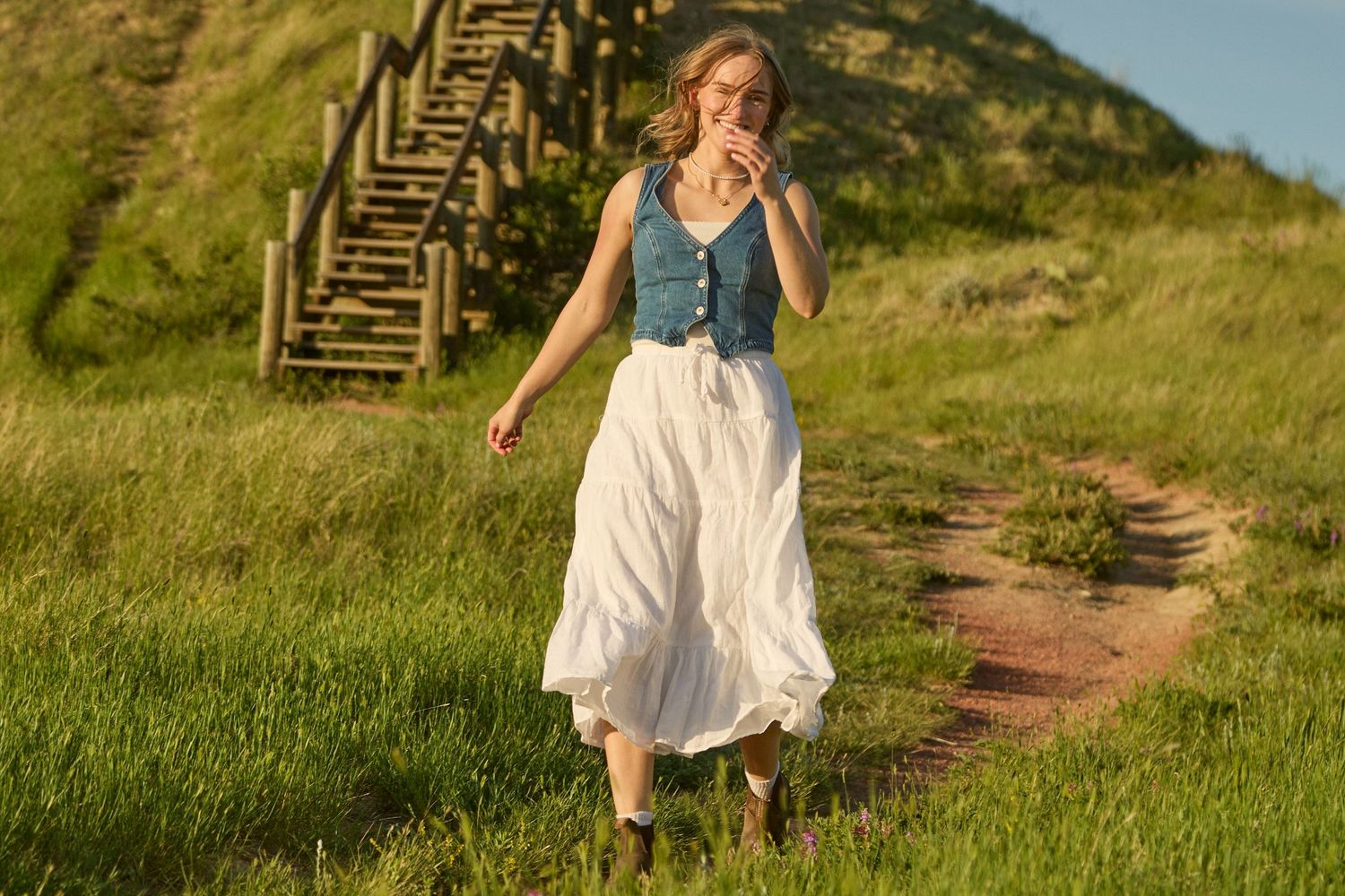 Young woman in denim vest and white maxi skirt walking on a scenic green hillside trail.