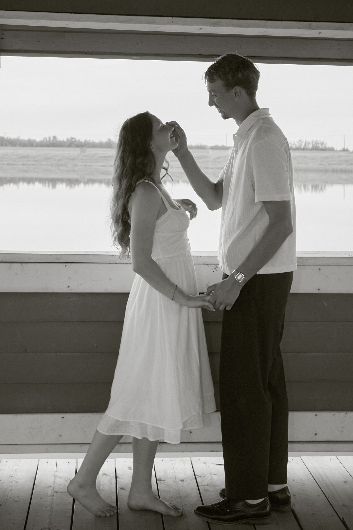 Black and white photo of a romantic couple holding hands by a lakeside window, woman in white dress.
