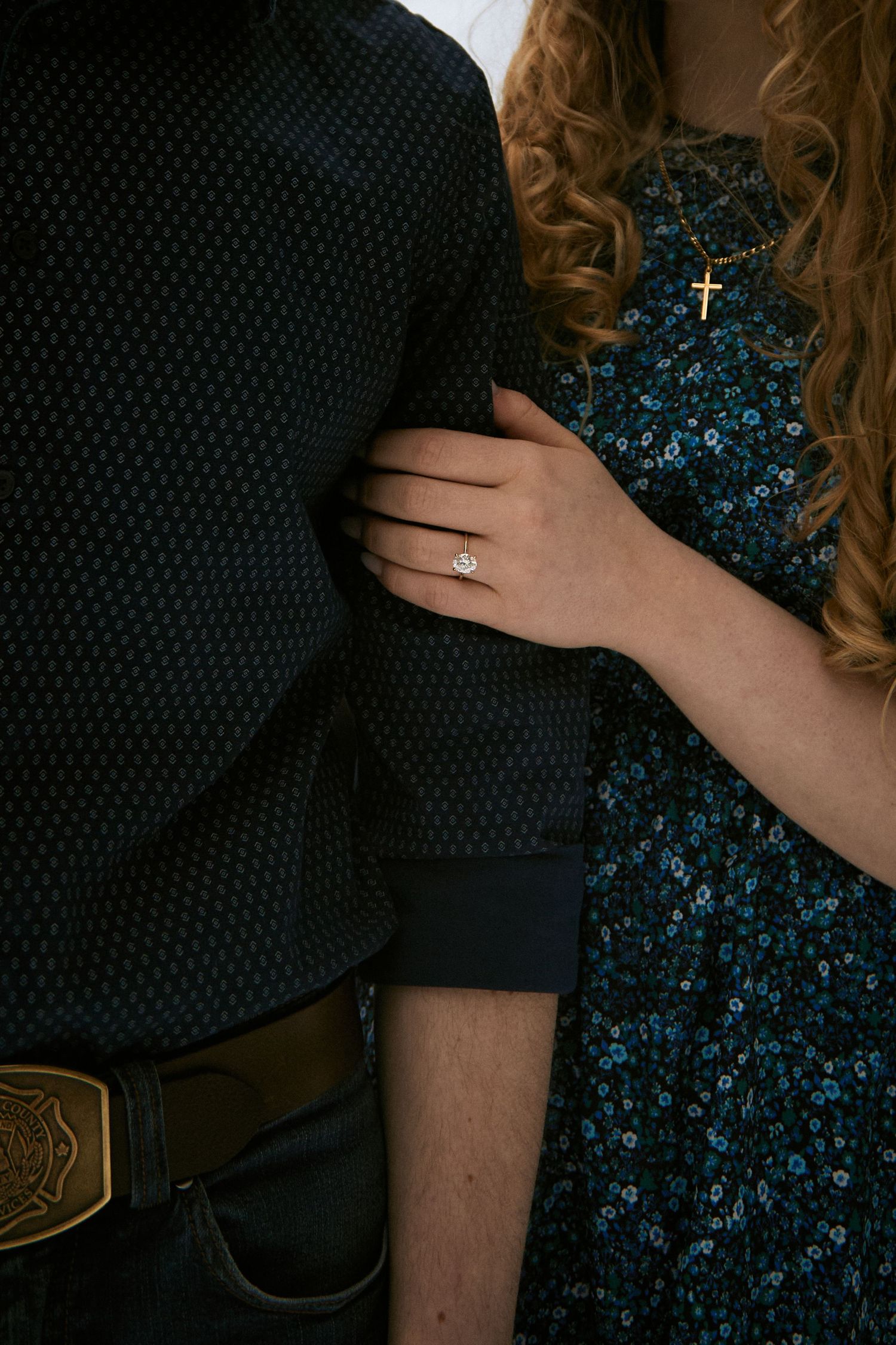 Couple embracing, woman wearing diamond engagement ring and gold cross necklace, man with firefighter belt buckle.