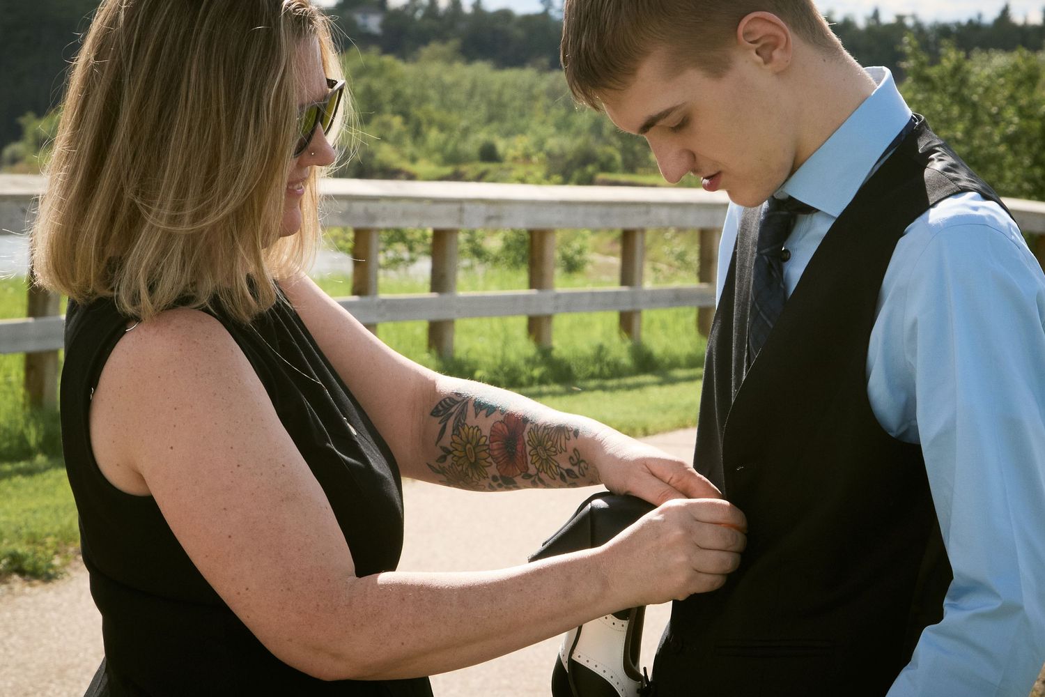 Woman with tattoo adjusting suit vest on young man in formal attire outdoors at scenic venue.
