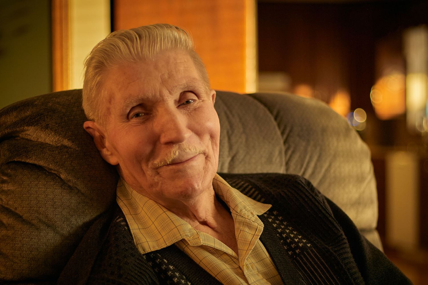 An elderly person smiles warmly while relaxing in a leather armchair with warm lighting in the background.