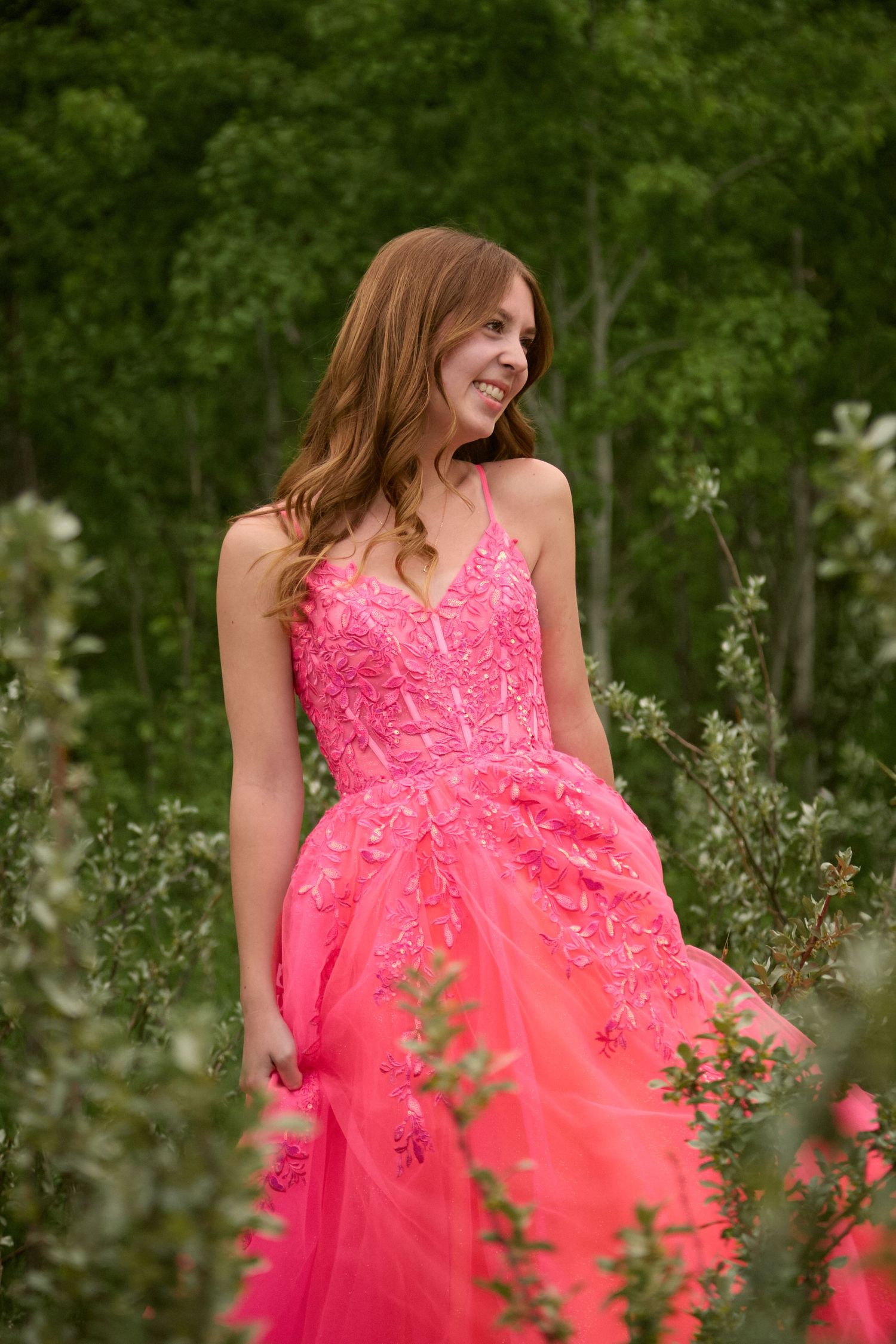 A smiling young woman in a bright pink floral lace ball gown poses outdoors among lush green shrubs and trees.