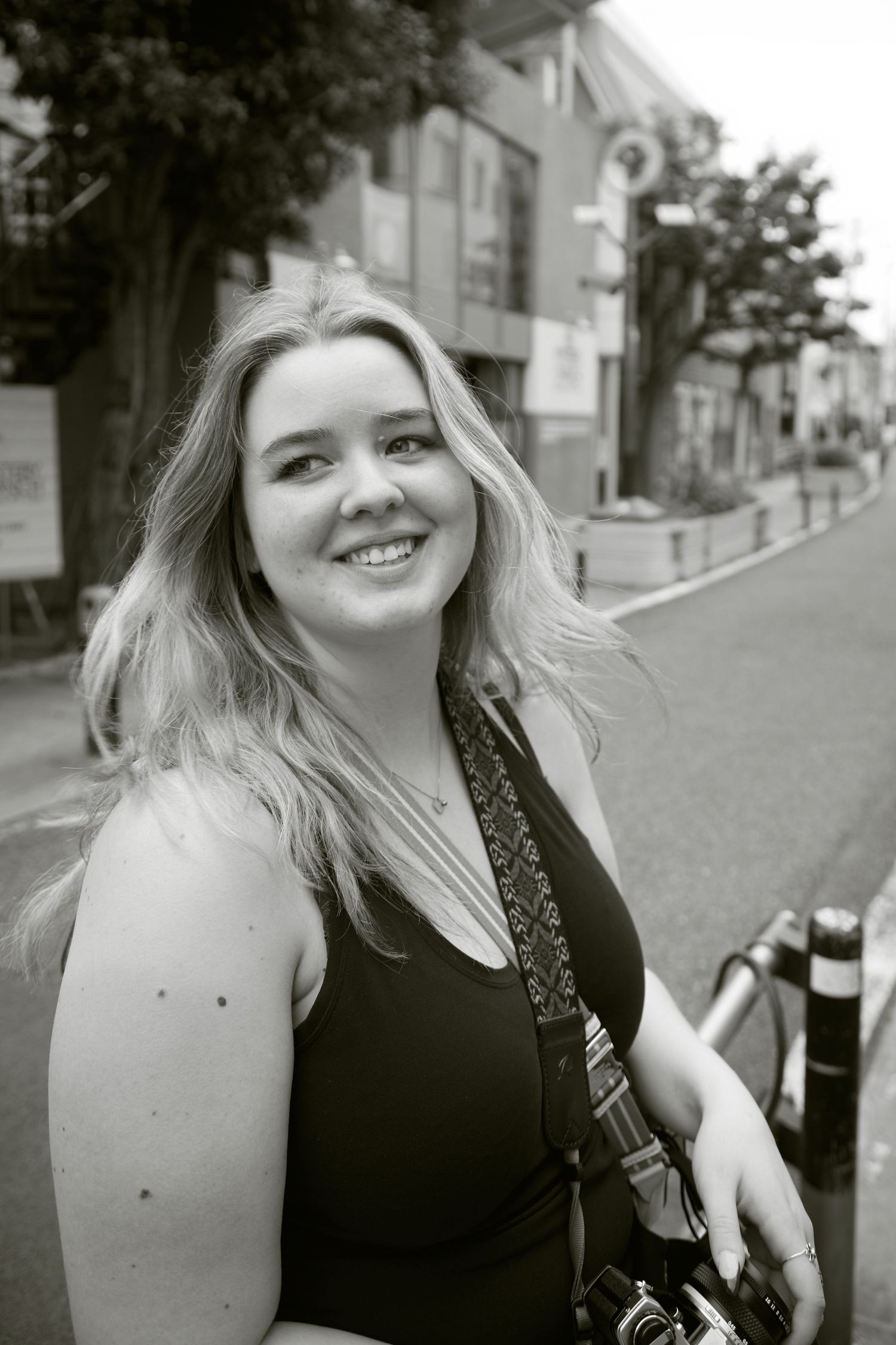Smiling blonde woman holding a camera on a city street in a black and white portrait photo.