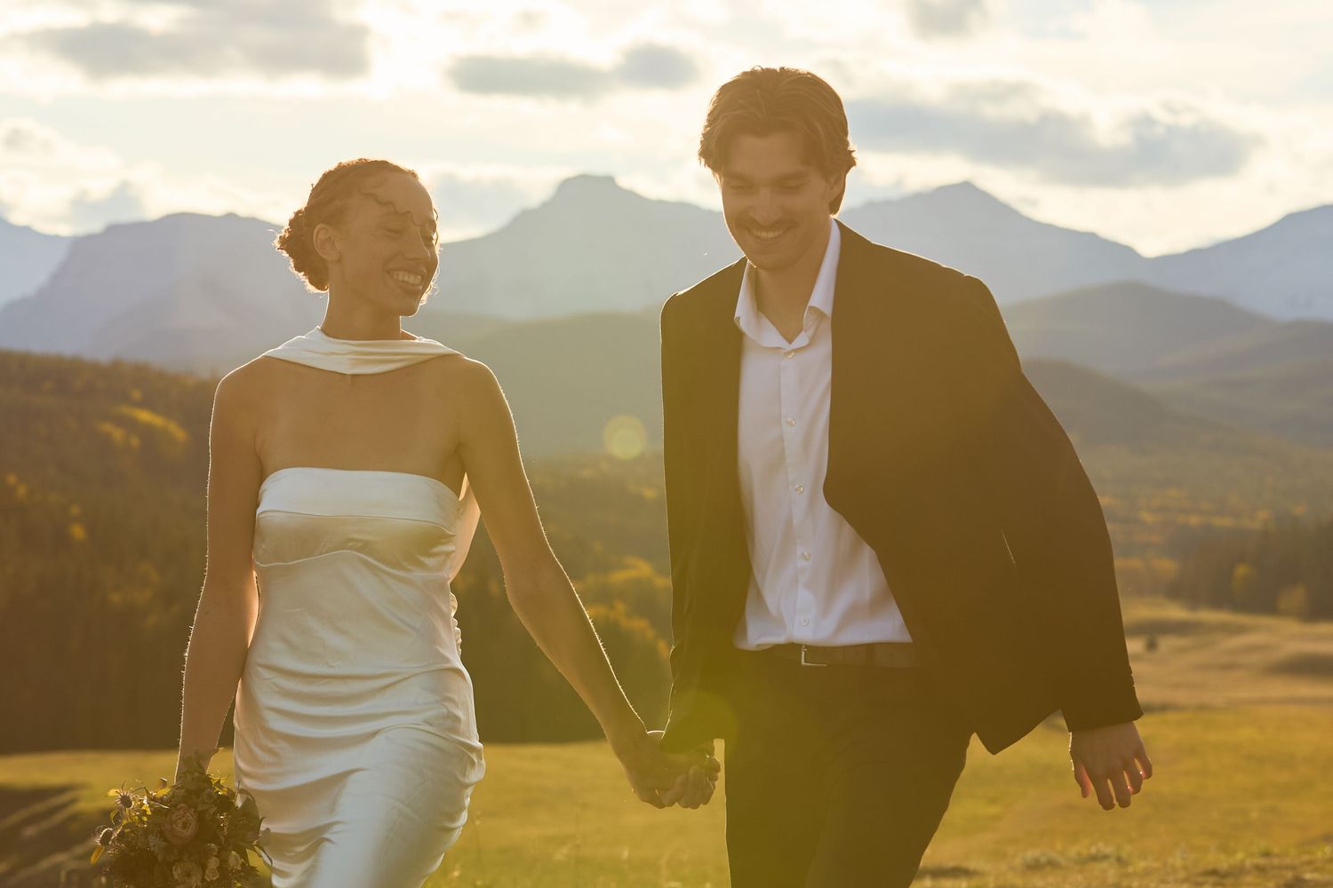 Couple embraces in a golden field at sunset with mountains in the background.