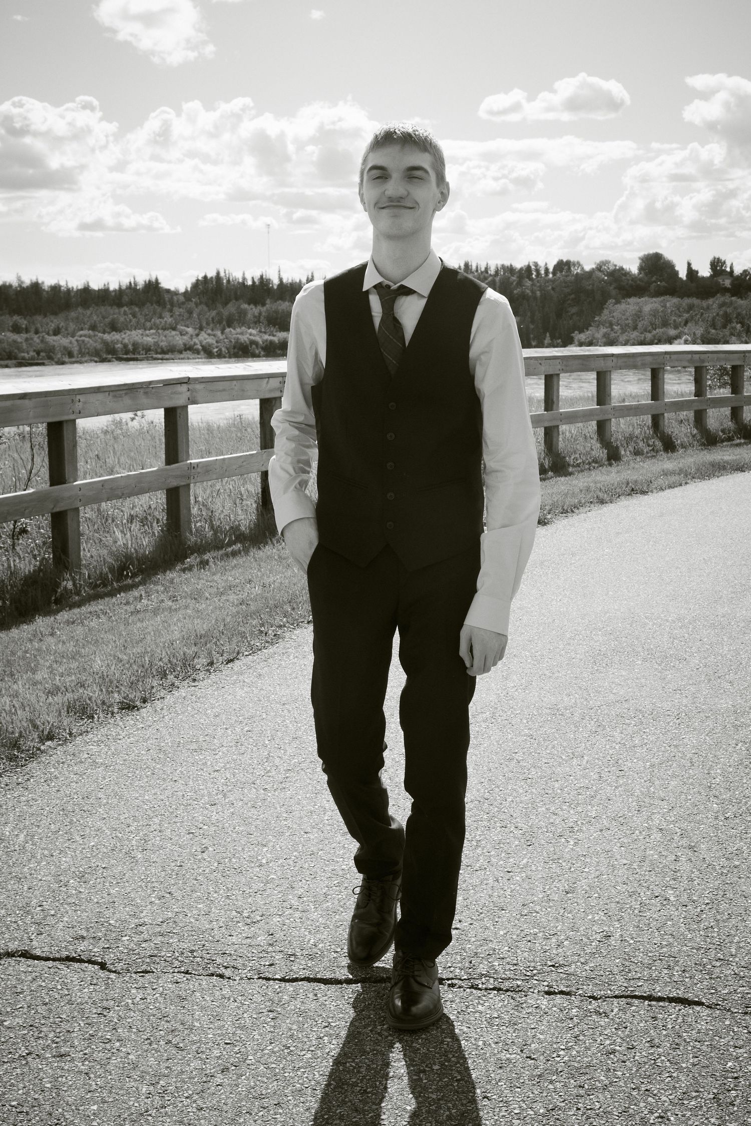 Black and white photo of a young man in a vest and tie walking on a path by a wooden fence and river.