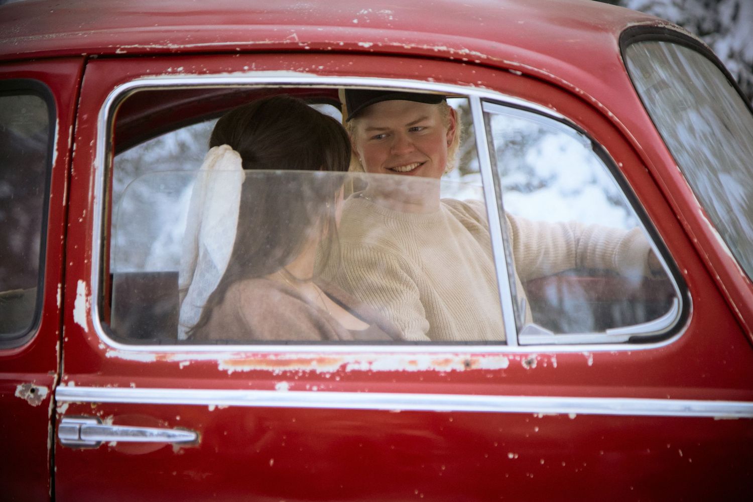 a couple smile inside a vintage red car, visible through the window.