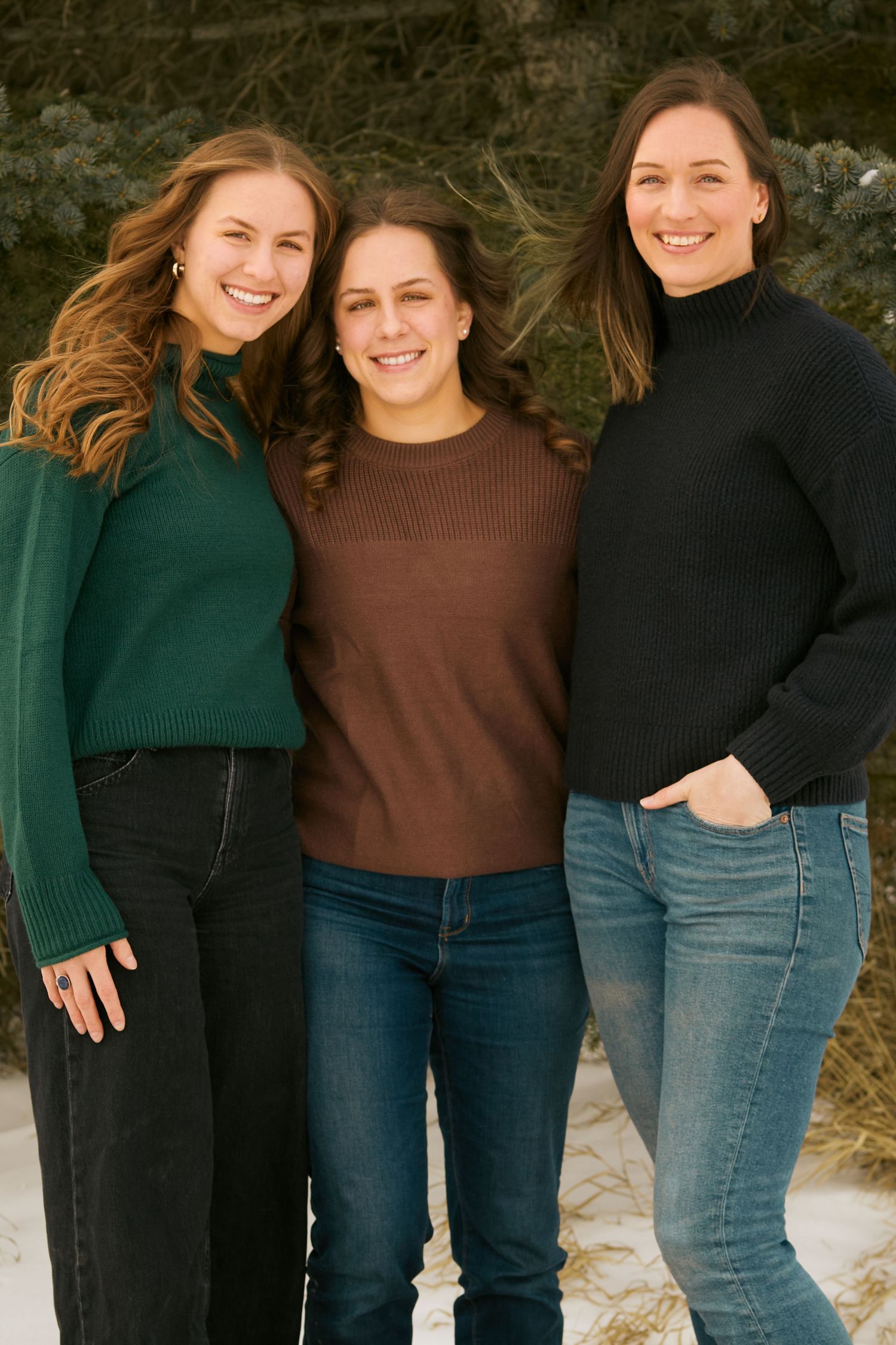 Three people wearing cozy sweaters and jeans pose together outdoors on a winter day.