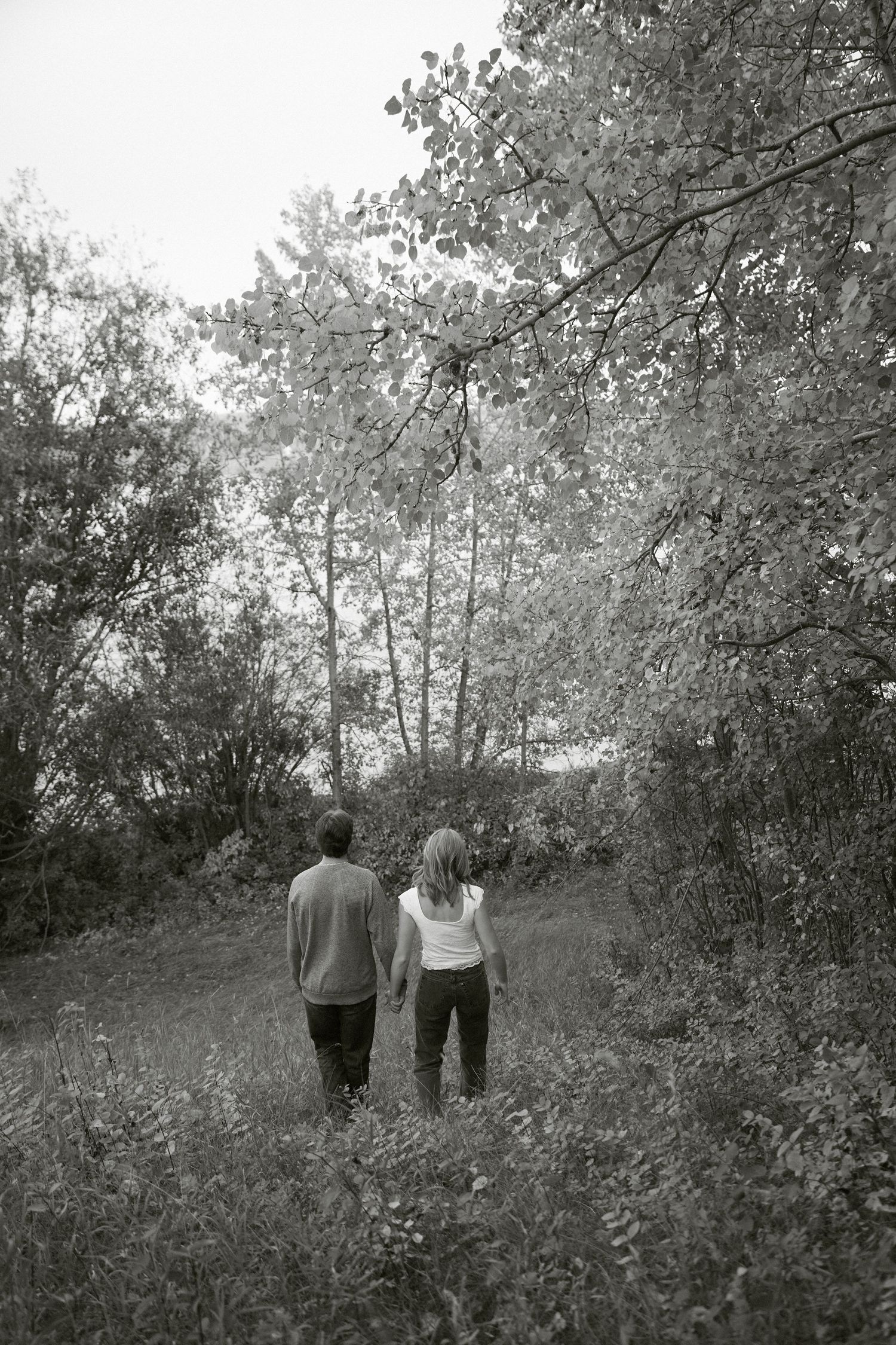 Black and white photo of a couple holding hands walking through a lush green forest trail.