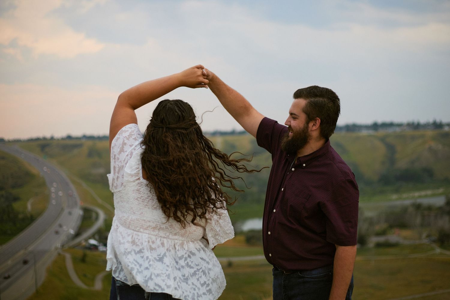 Couple dancing outdoors on a hilltop at sunset with a scenic highway and green valley in the background.