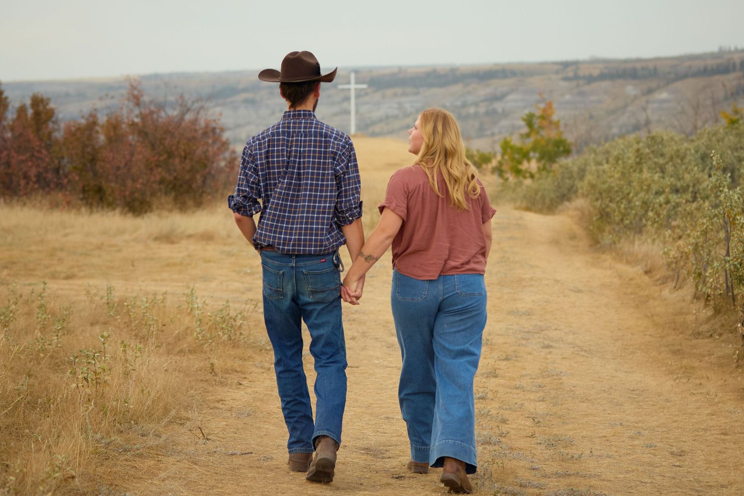 A couple walking hand in hand on a dirt trail through autumn hills wearing casual western attire.
