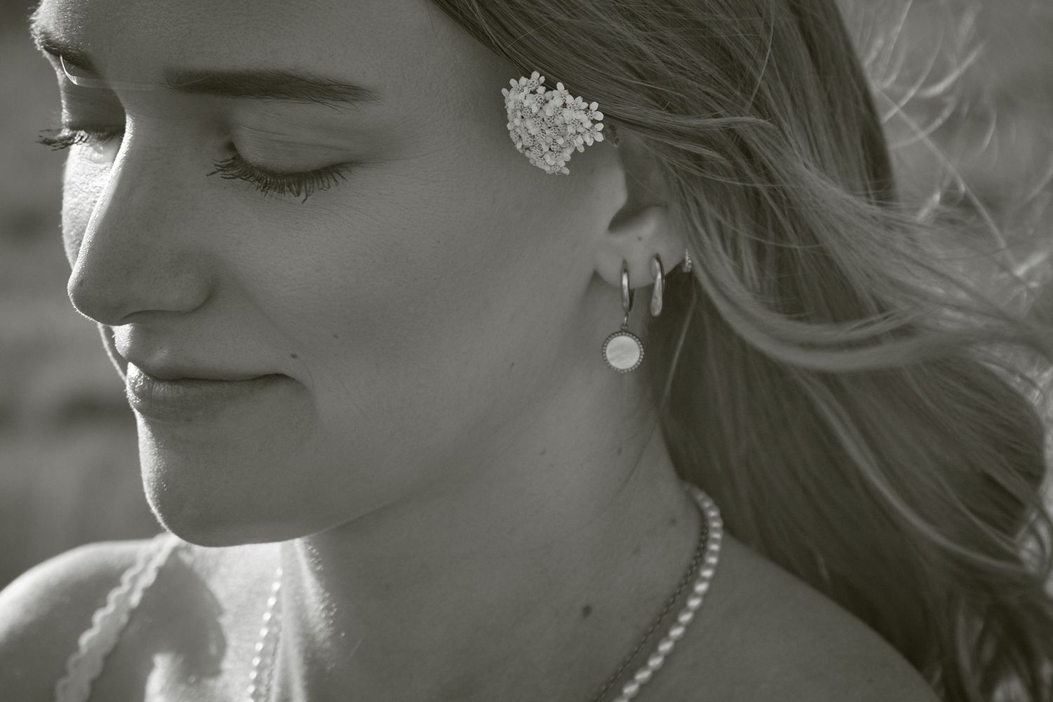 Black and white close-up of a bride wearing pearl necklace, hoop earring, and floral hair accessory.