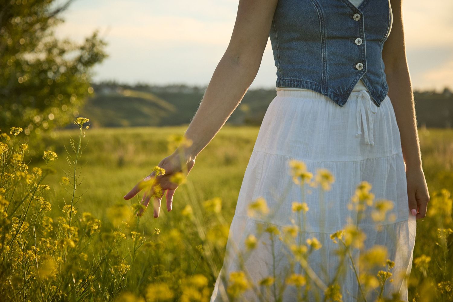 Woman in white skirt and denim vest walking through a sunny yellow wildflower field at golden hour.