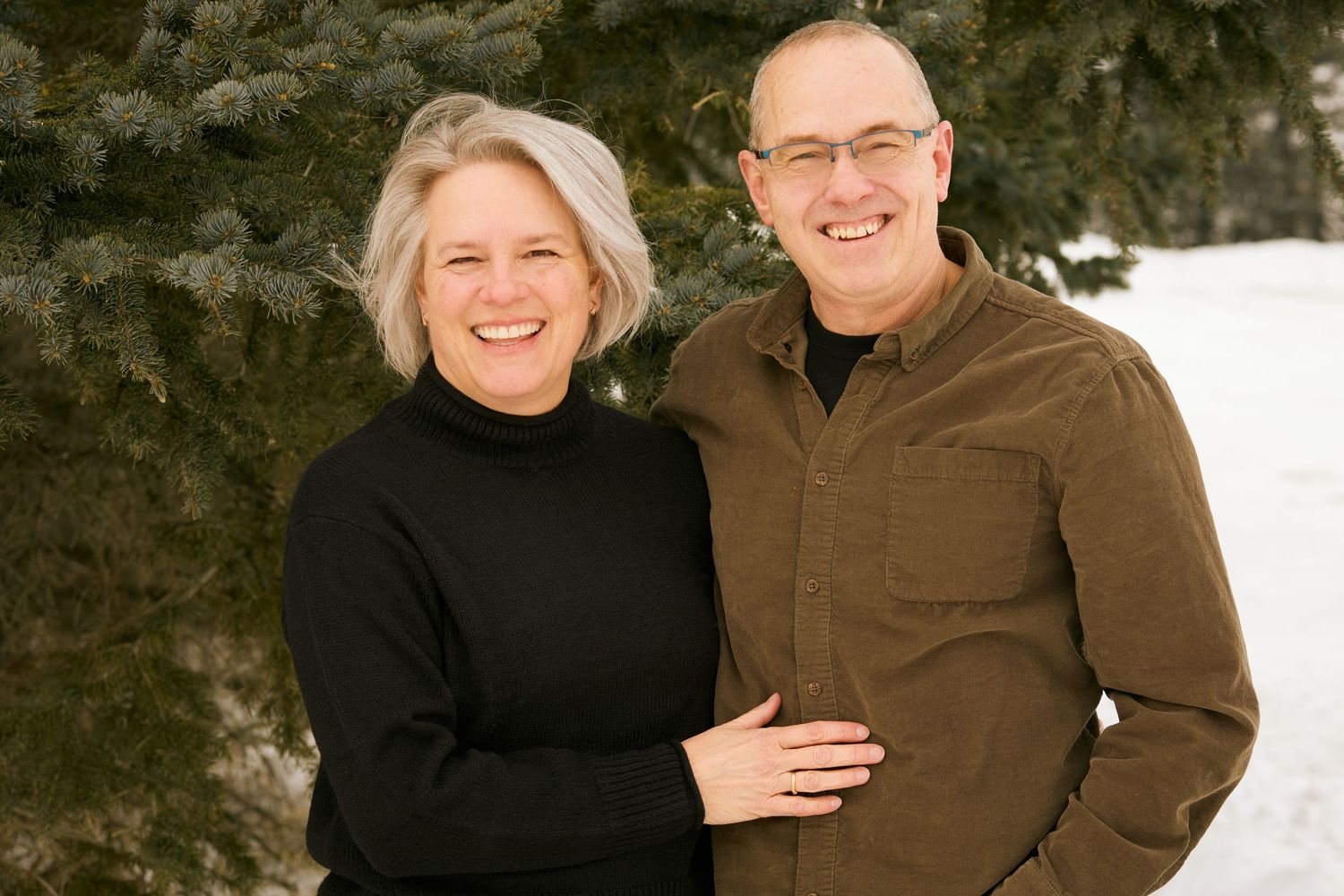 A couple in black and brown winter attire share warm embraces while posing outdoors in the snow.