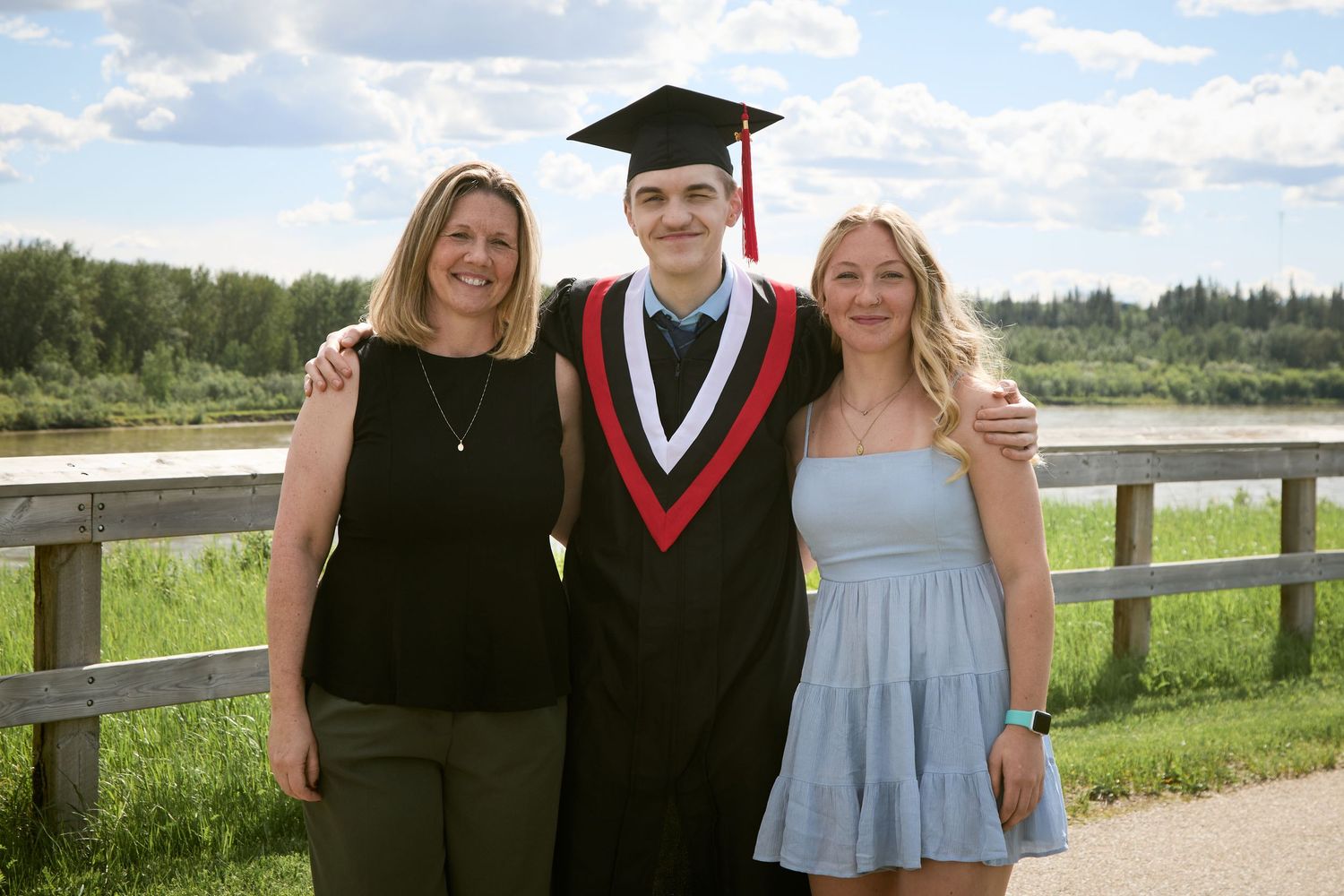 Graduate in cap and gown poses with two smiling women outdoors near a wooden fence and lush green landscape.