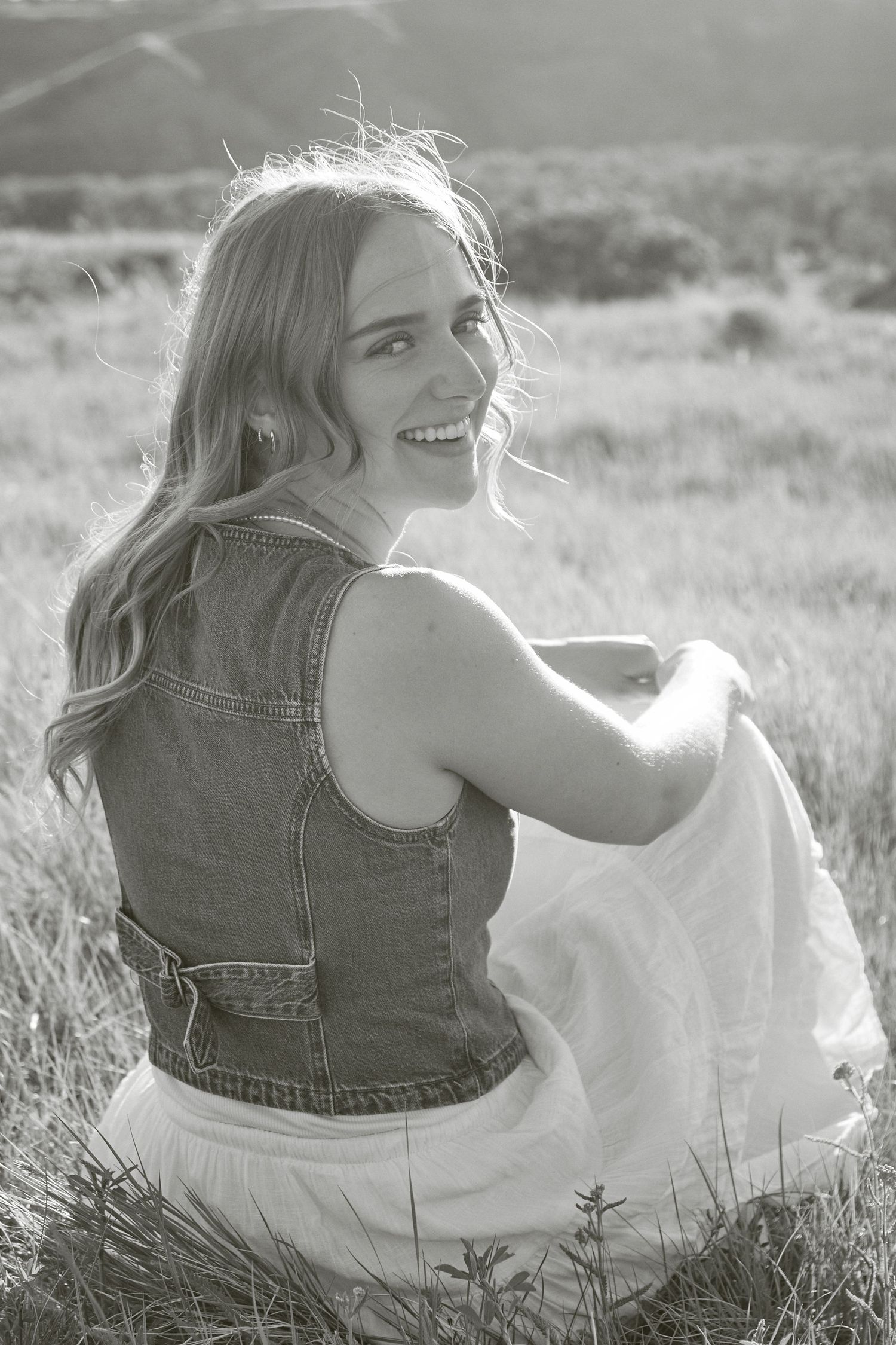 Smiling blonde woman in denim vest and white skirt sitting in a sunlit field, captured in black and white.