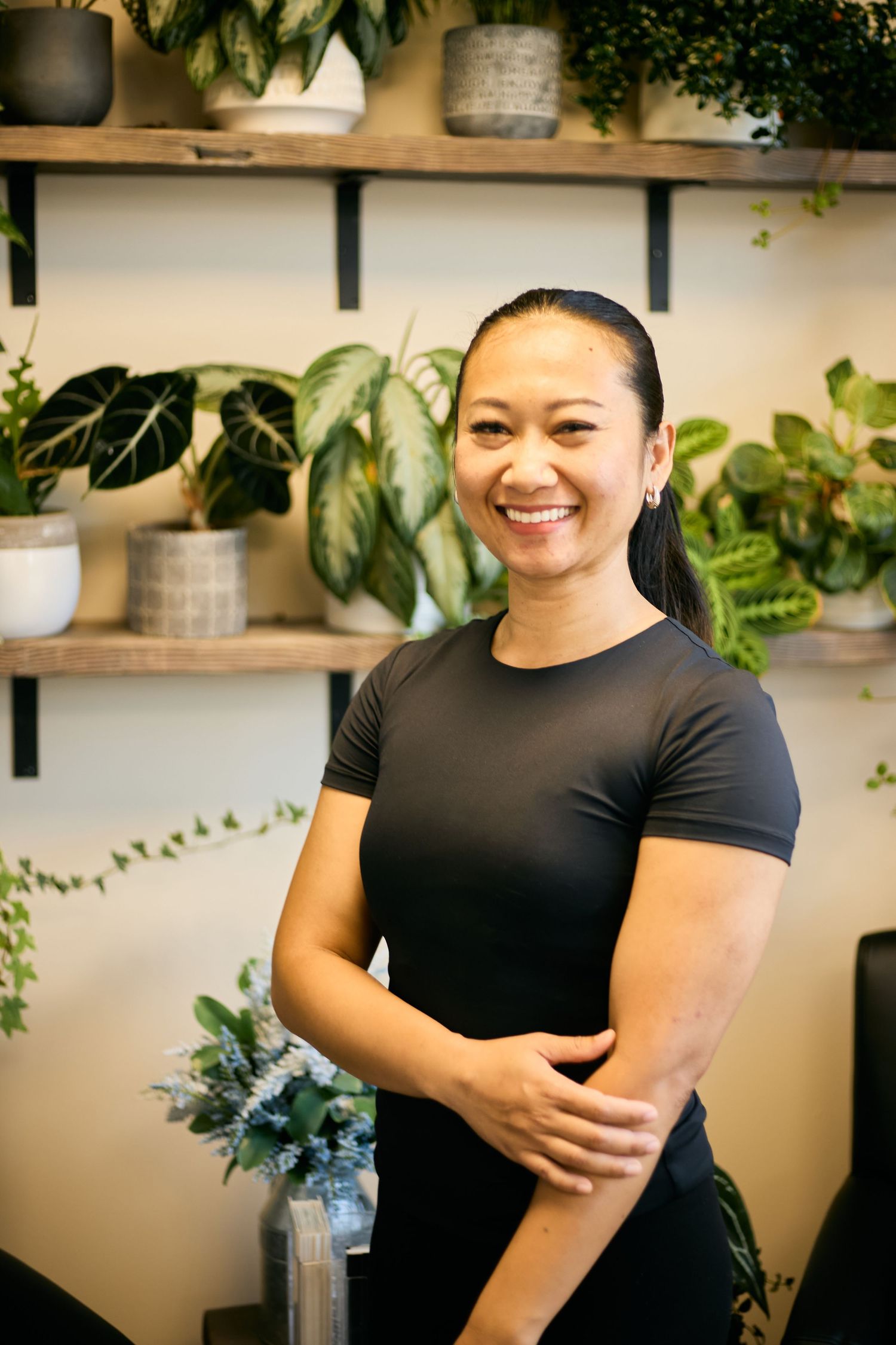 Massage therapist in black uniform standing in spa room with plants on shelves.