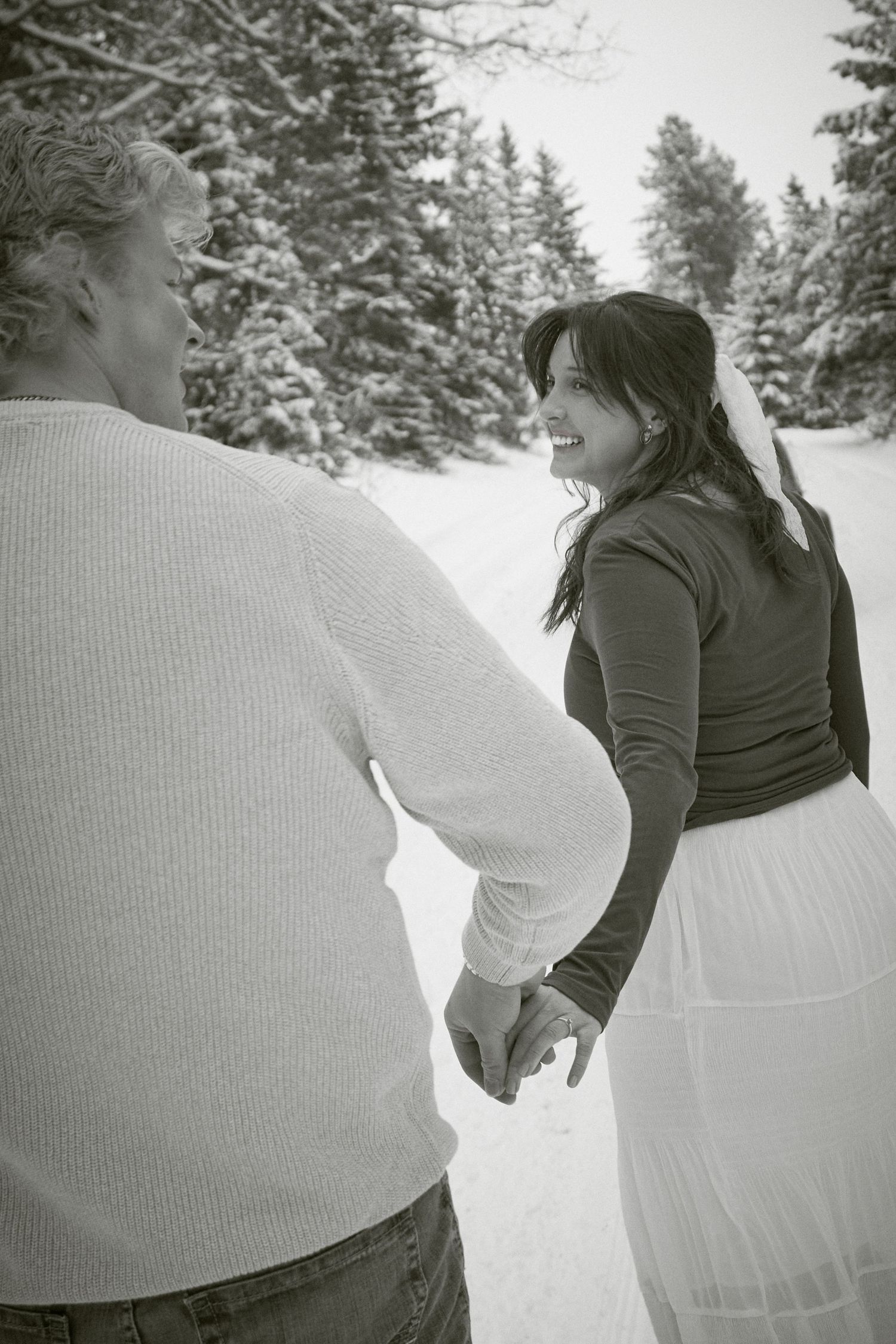 Black and white photo of a smiling bride holding hands with her partner in a snowy winter landscape.