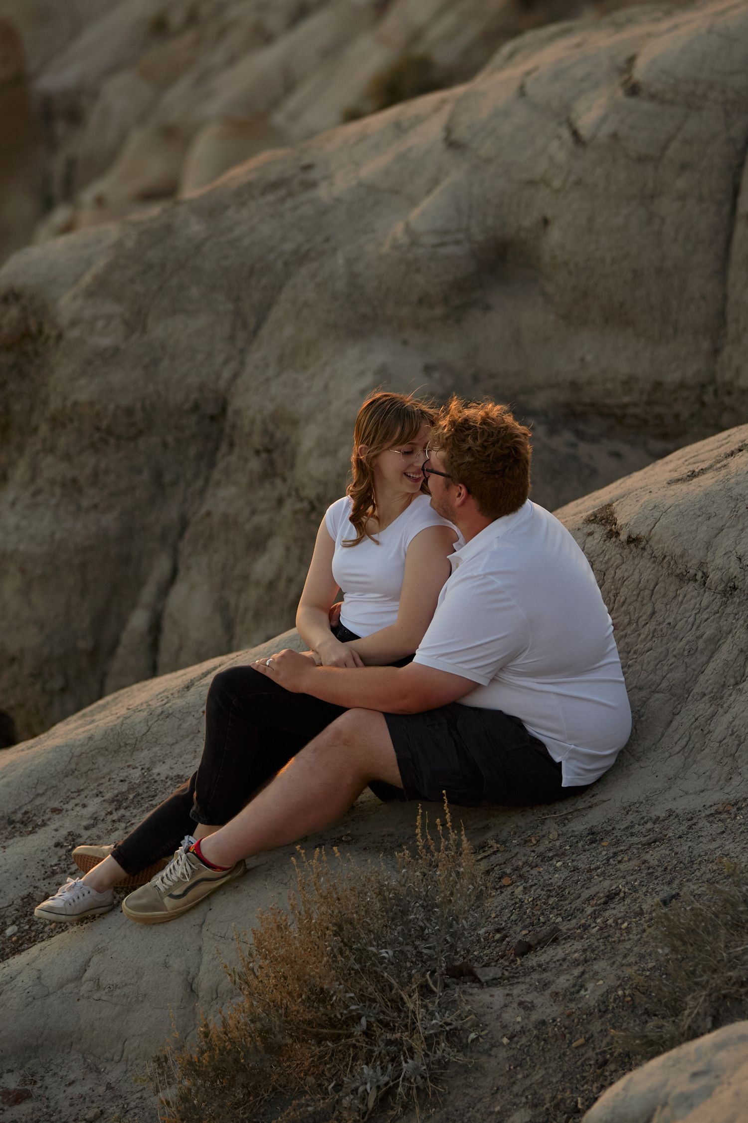 Couple in white shirts sitting on rocky terrain, sharing a romantic moment at golden hour.